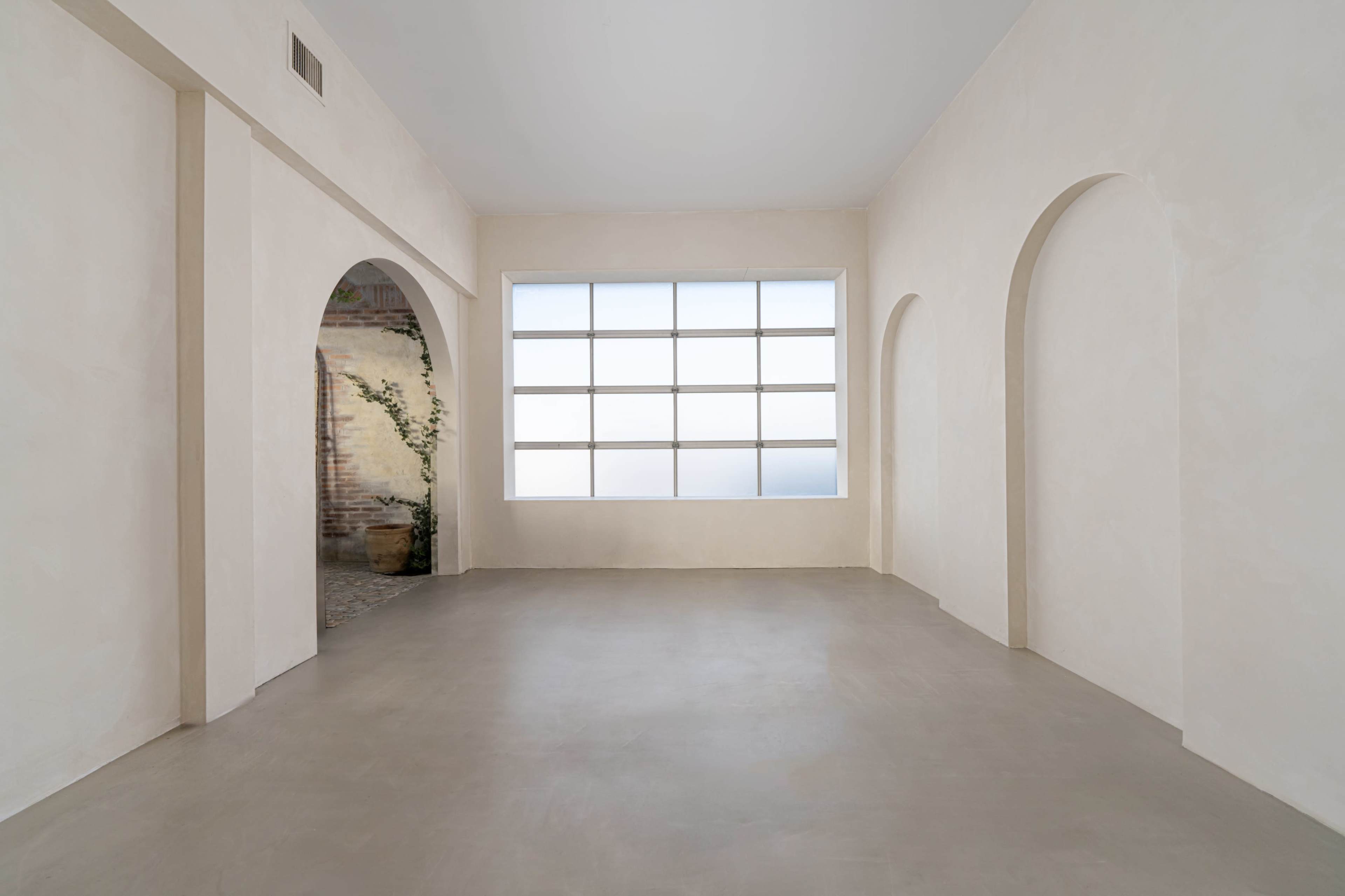 A spacious interior with plain walls, a large window, and two arched openings leading to another area.