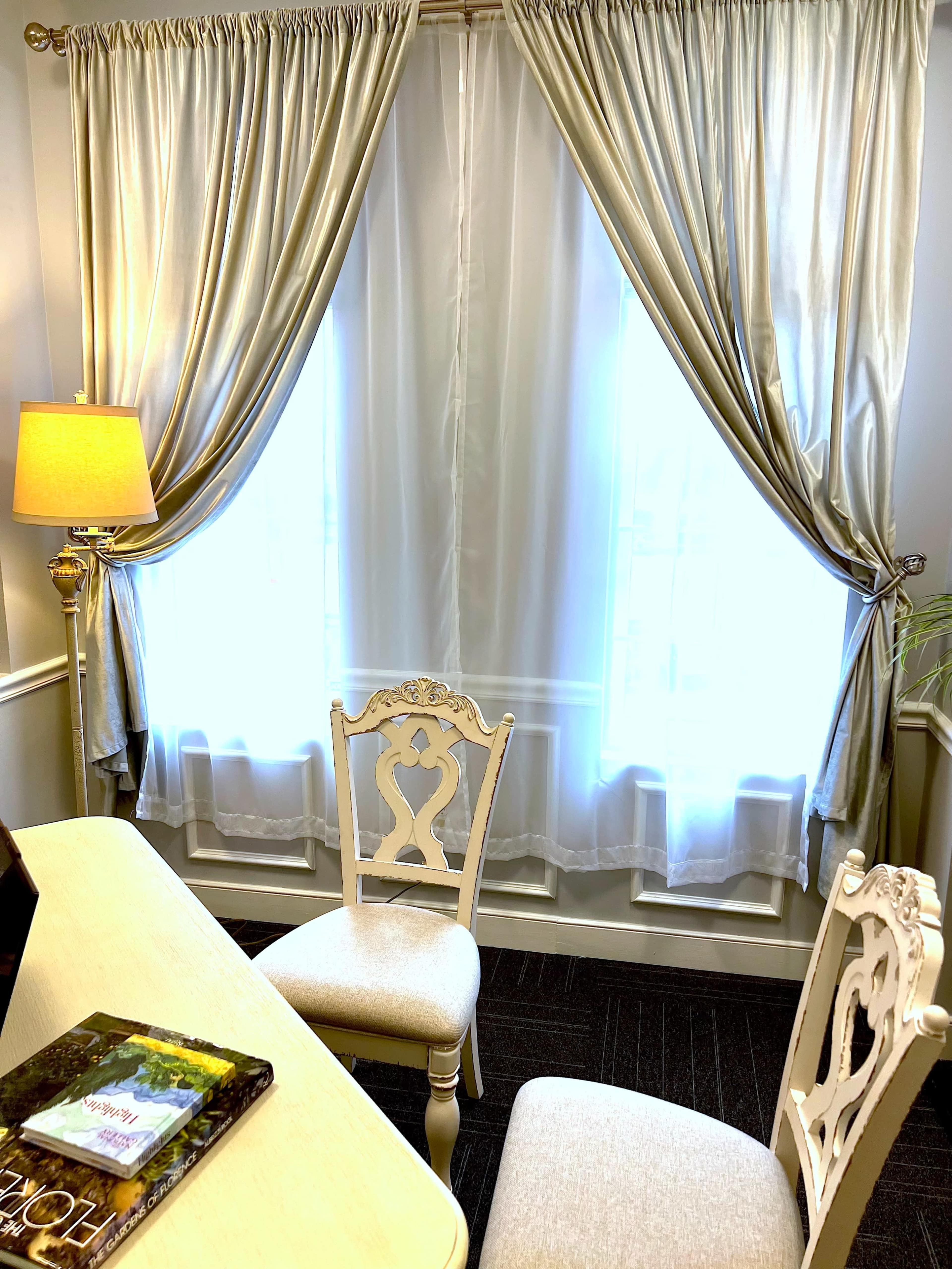 The image shows a well-lit room with sheer curtains, a lamp, a light-colored desk, and two ornate chairs.