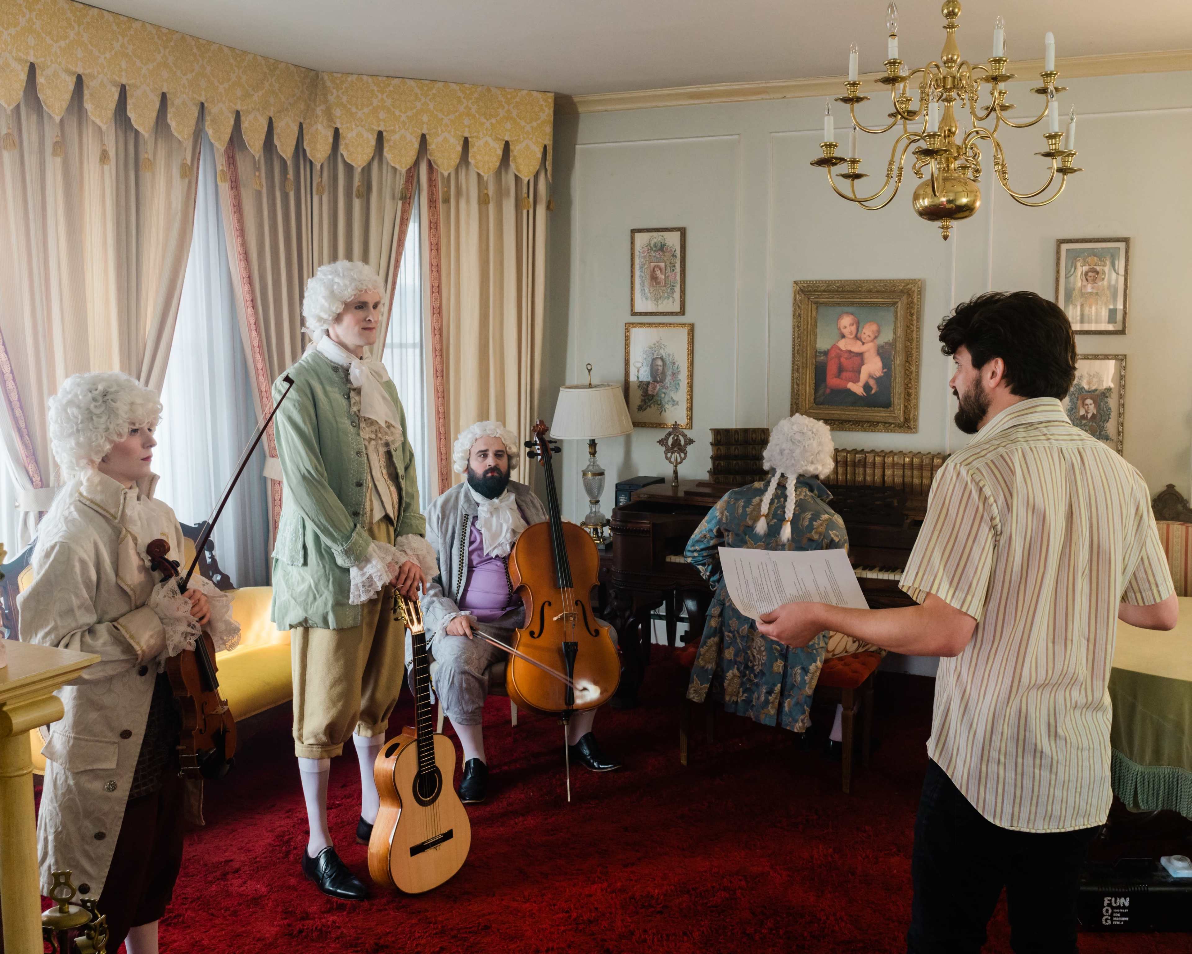 A group of musicians dressed in period costumes plays instruments in a styled room while a man stands holding a script.