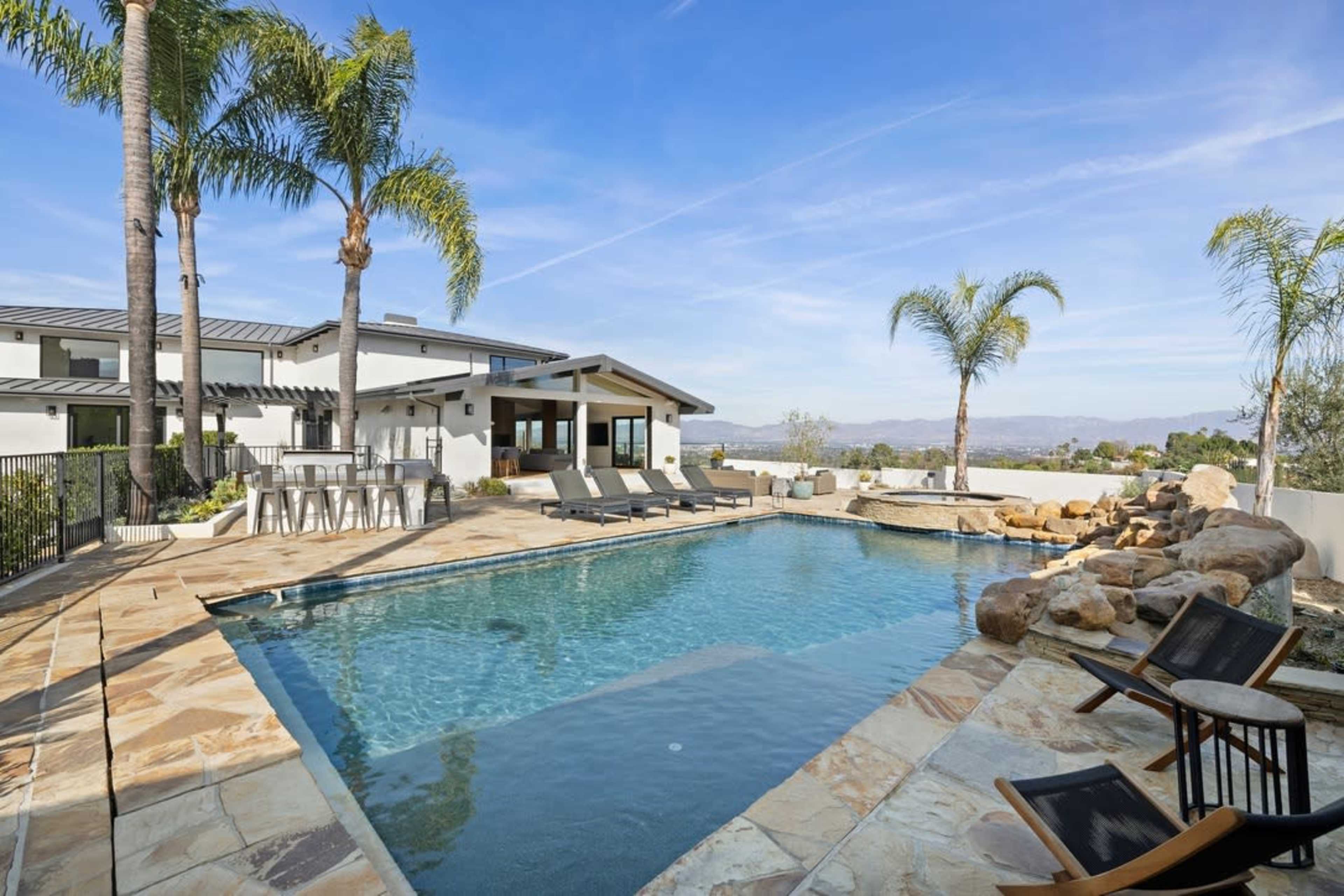 The image shows a modern outdoor pool area with palm trees, lounge chairs, and a scenic view of the hills in the background.