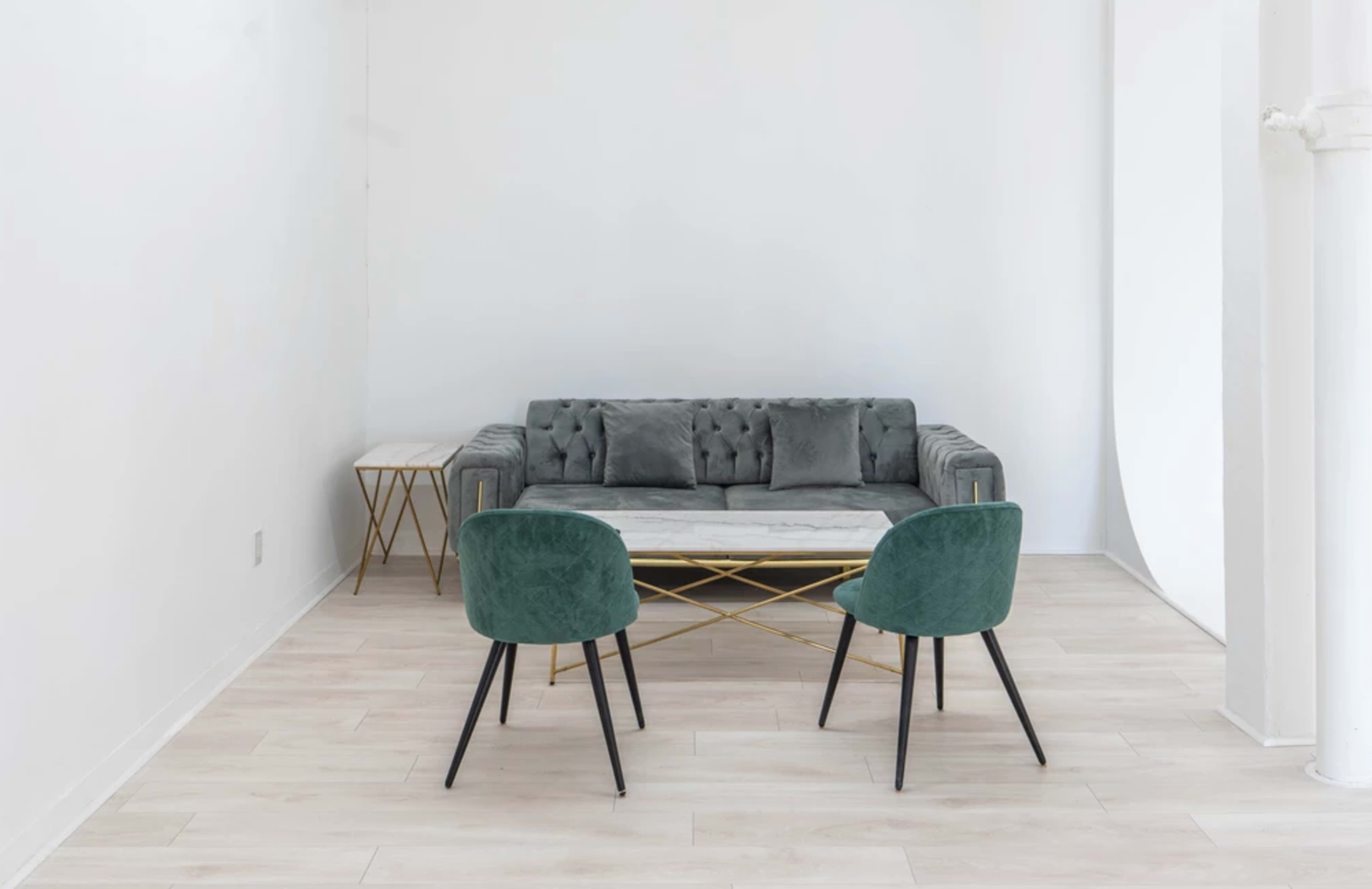 A modern living space features a gray tufted sofa, a marble-topped coffee table with gold legs, and two green upholstered chairs on a light wood floor.
