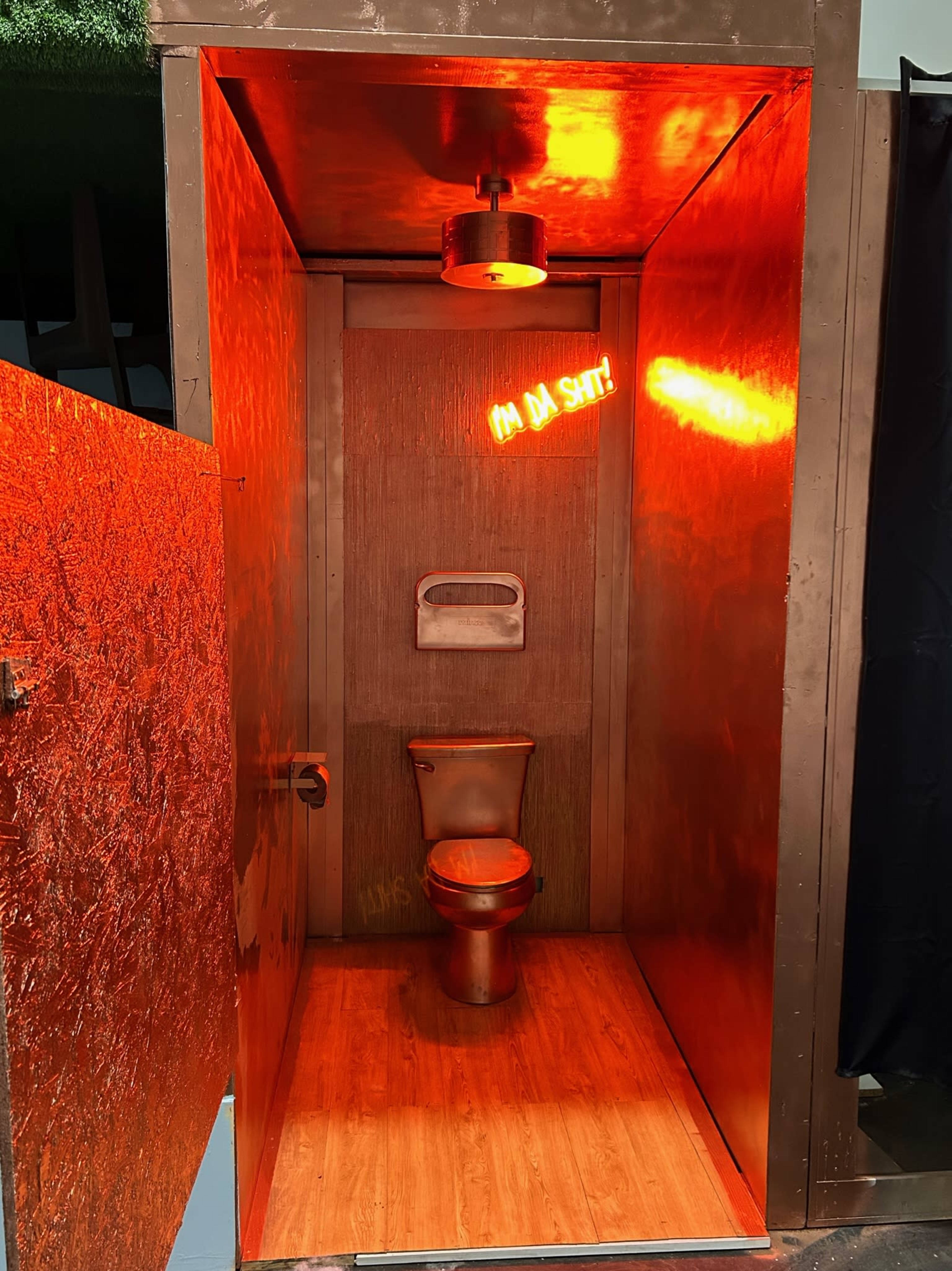 A small, metallic-walled restroom stall with a toilet and a glowing neon sign that reads "I'M DA SH*T."