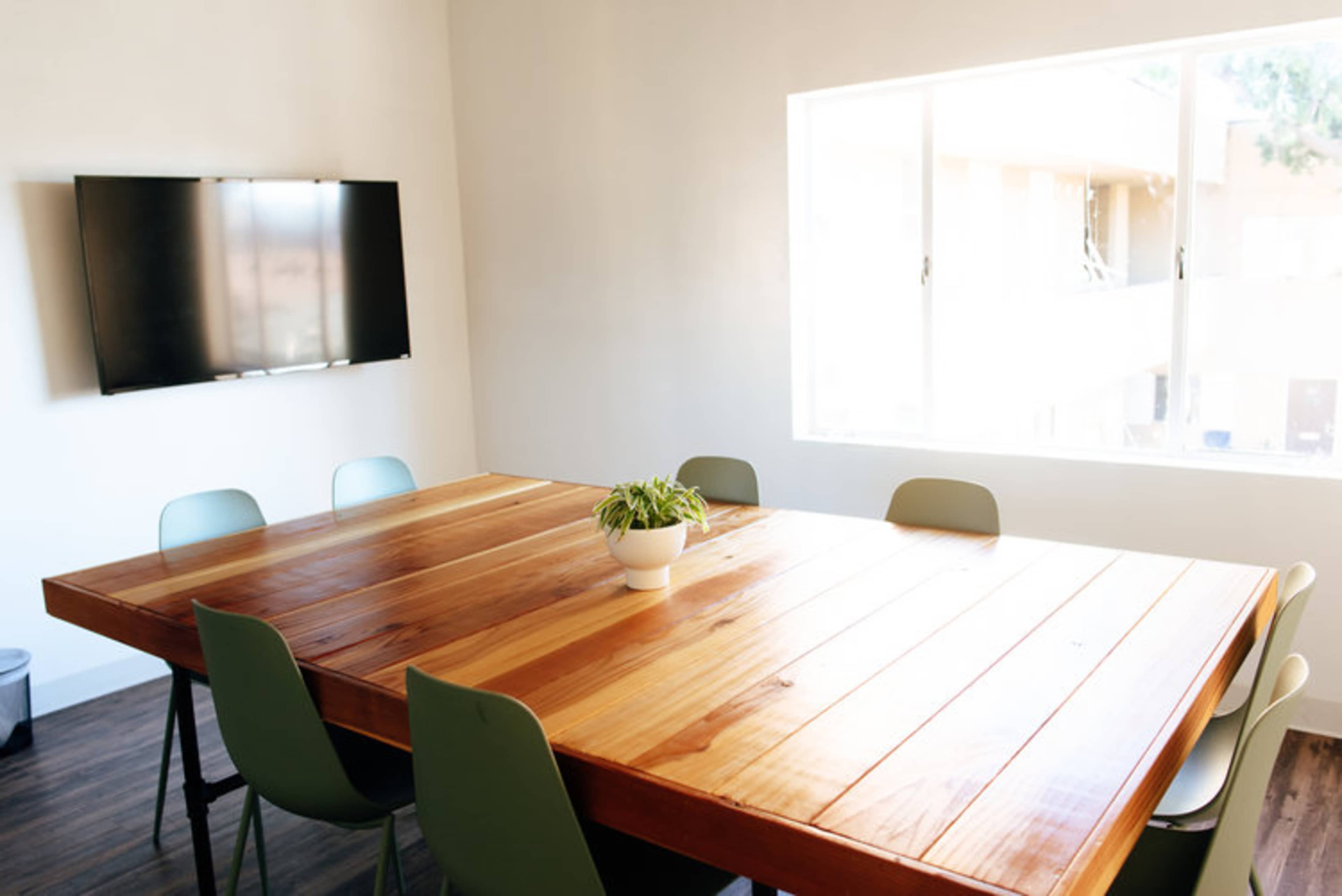 A large wooden table surrounded by chairs is positioned in a brightly lit room with a television mounted on the wall and a window offering an outside view.