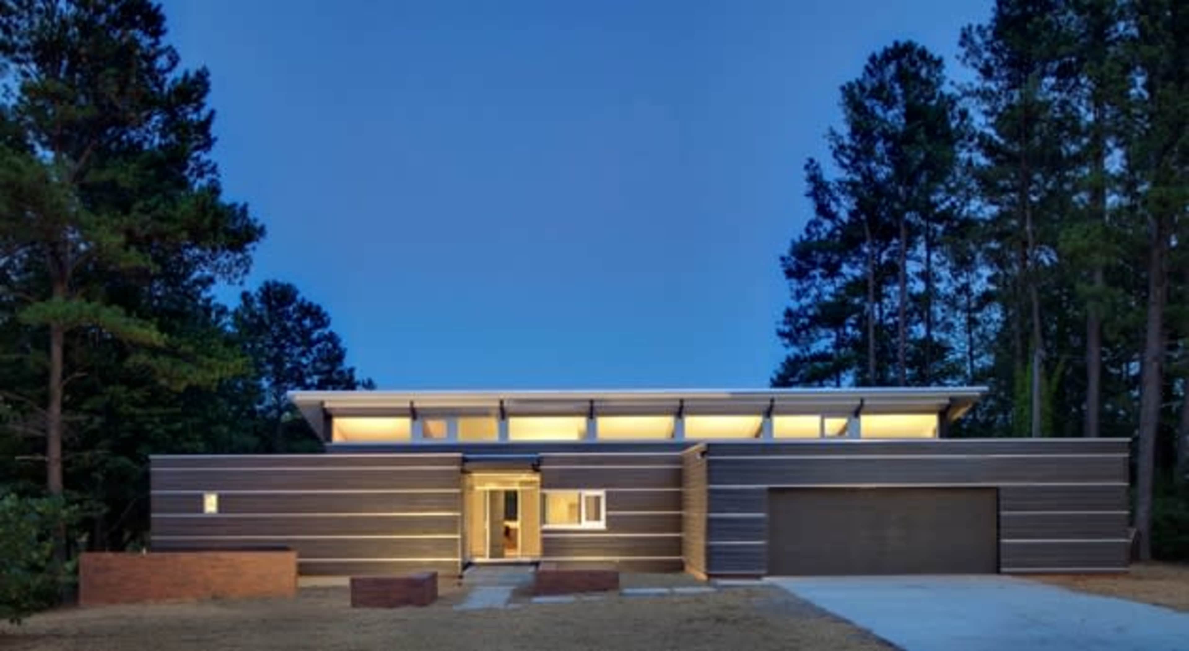 A modern house with horizontal wood paneling and large windows is set against a backdrop of tall trees at dusk.