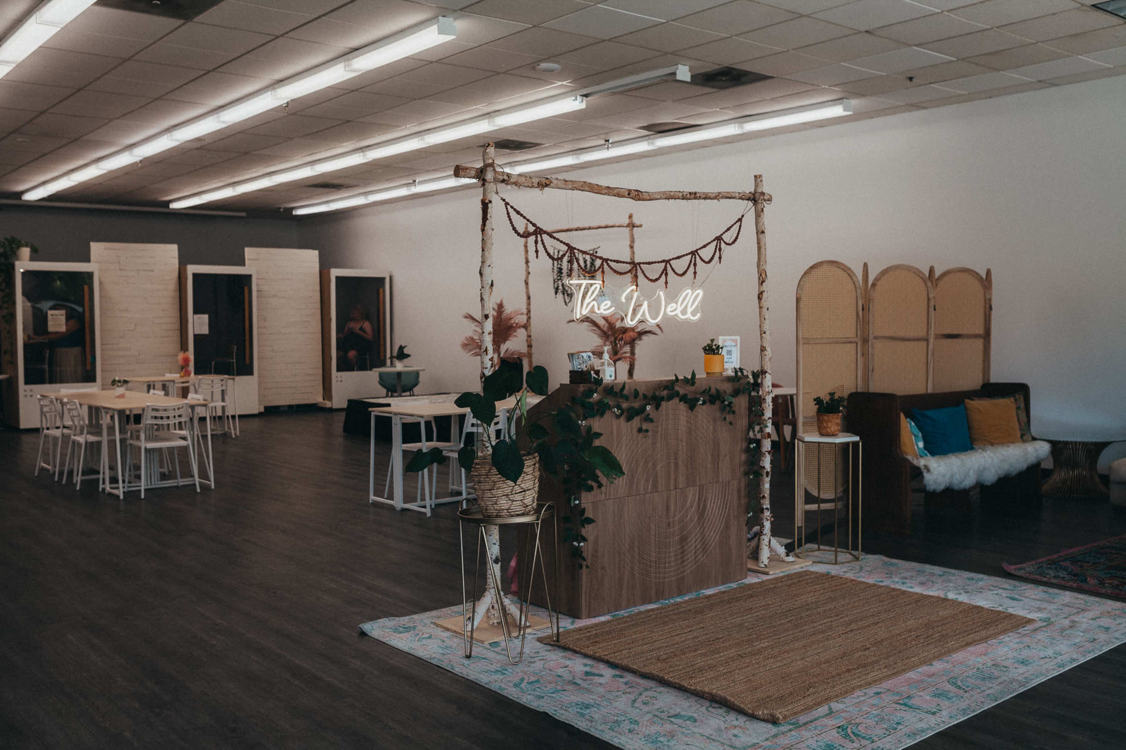 The image shows a light and airy interior with a wooden reception area labeled "The Well," surrounded by tables and chairs in a spacious room.