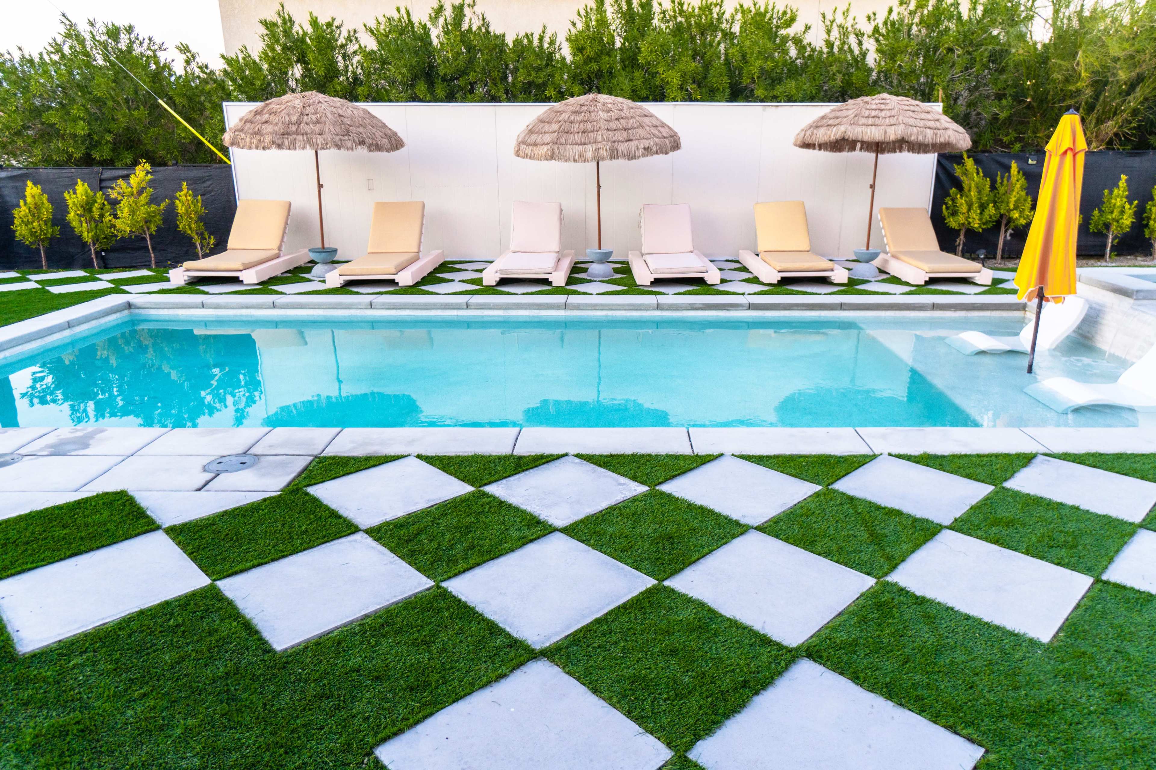 A swimming pool surrounded by lounge chairs, straw umbrellas, and a checkerboard-patterned lawn.