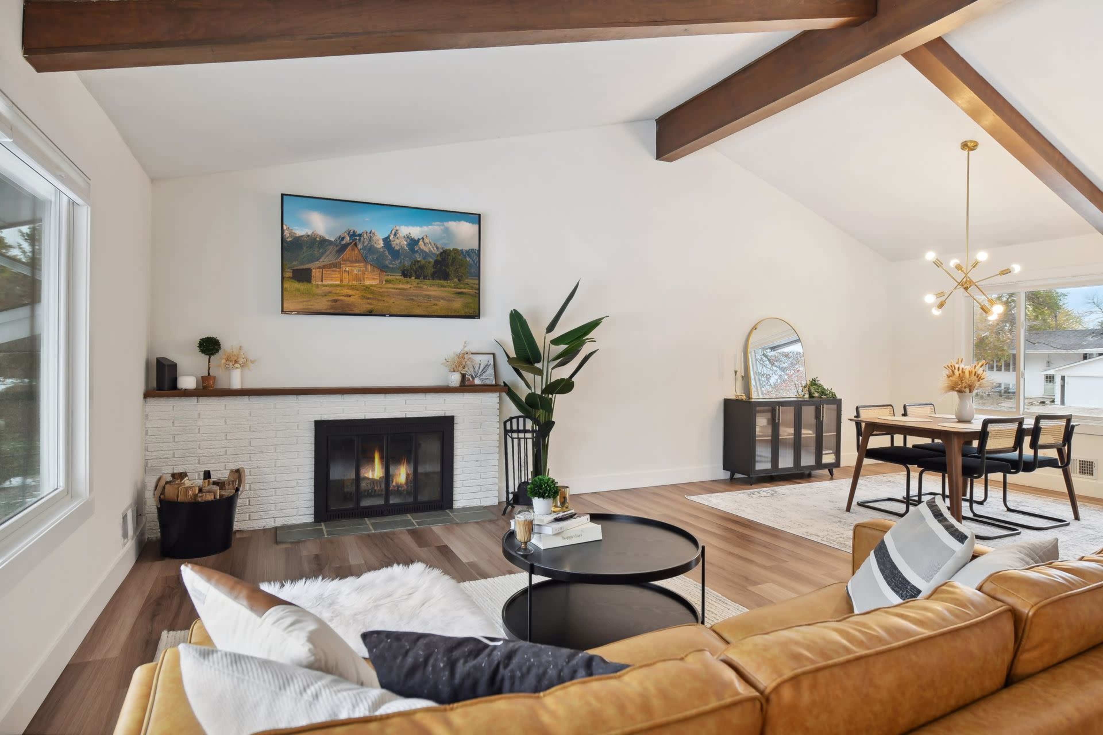 The living room features a cozy seating area with a leather sofa, a coffee table, and a fireplace beneath a large mountain landscape painting, accompanied by a dining space with a round table and modern light fixture.