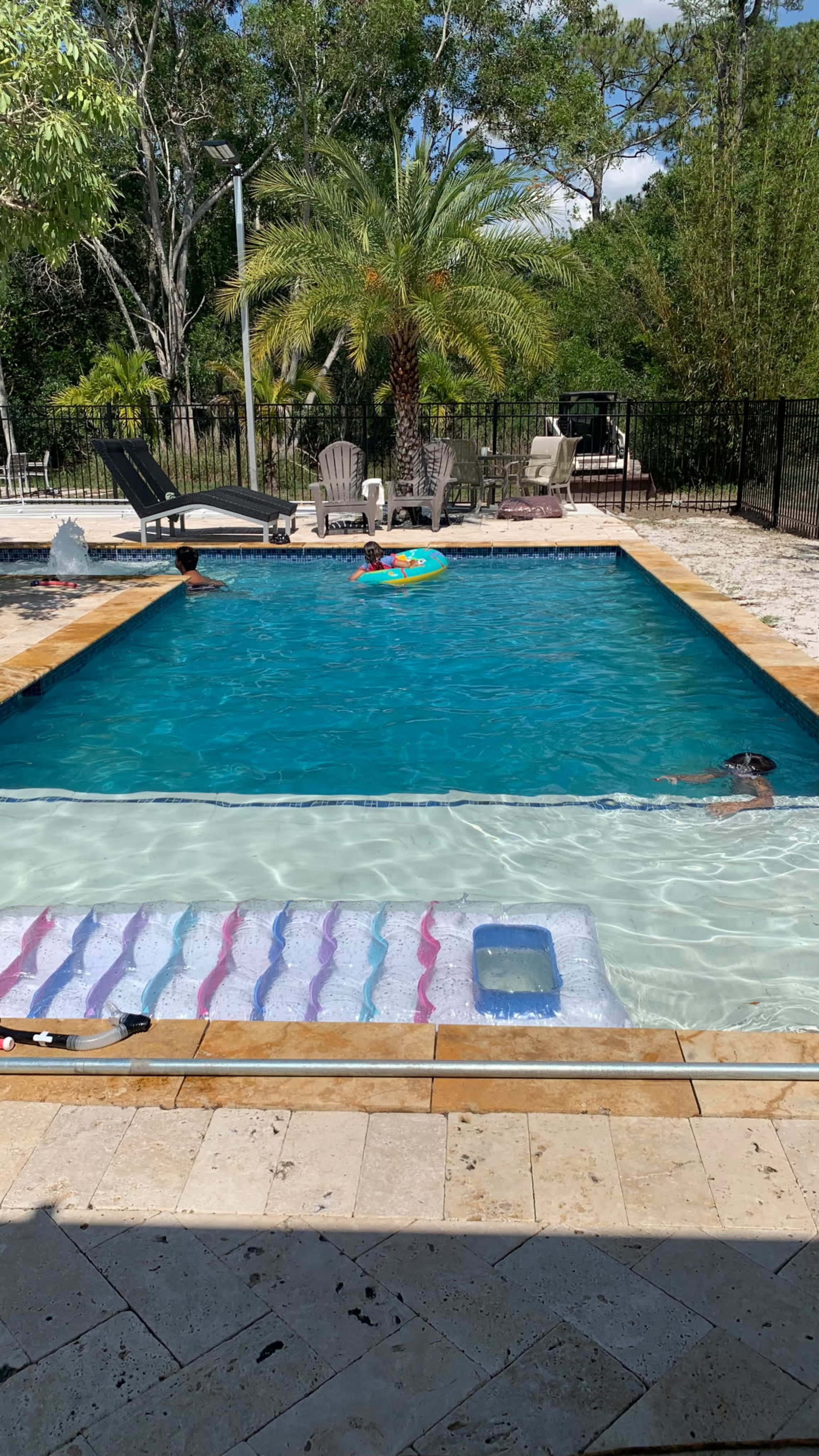 A swimming pool is surrounded by chairs and palm trees, with people floating on inflatables in the water.