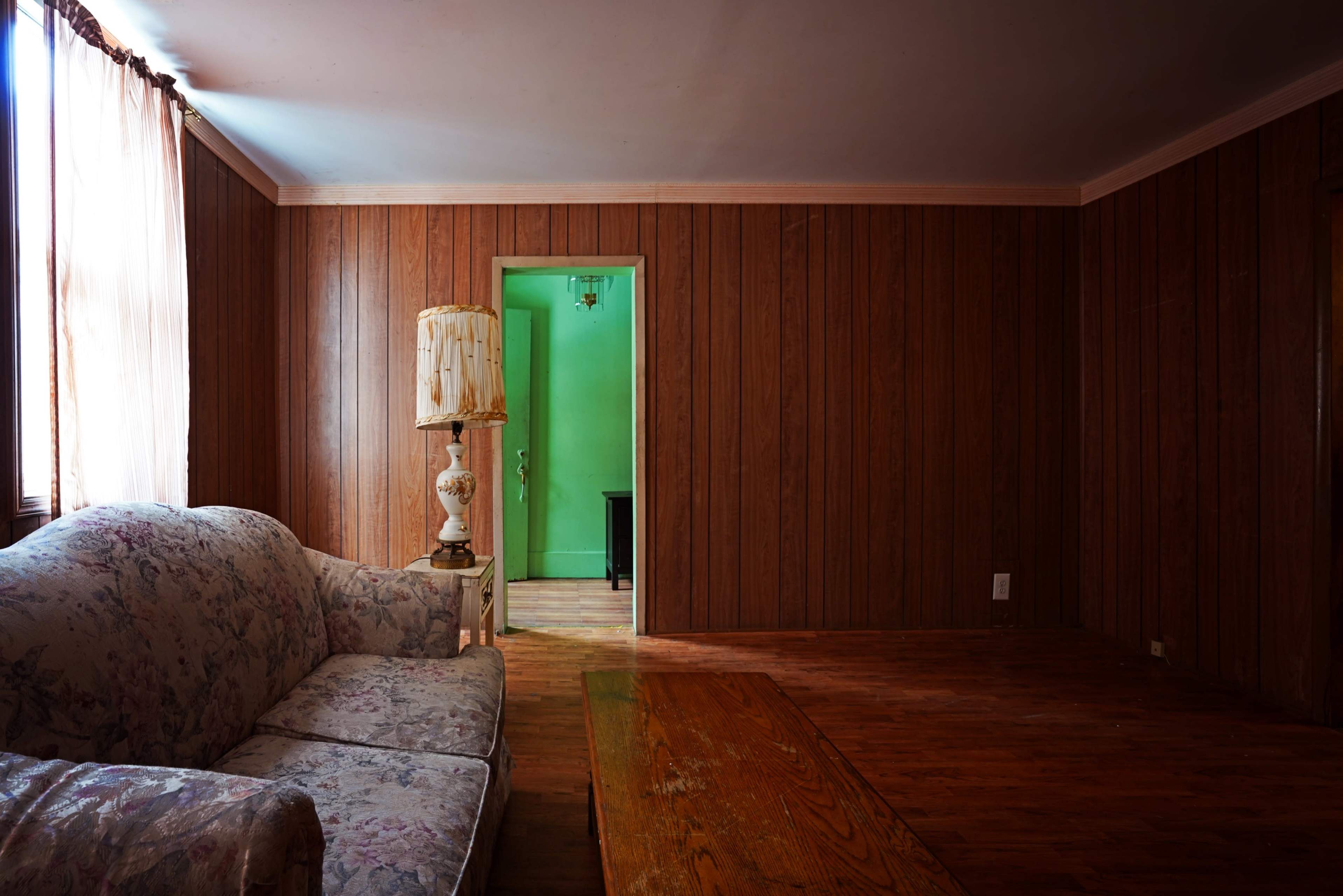 A room with wooden paneling, a floral-patterned couch, a wooden coffee table, and a doorway leading to a green-lit space.