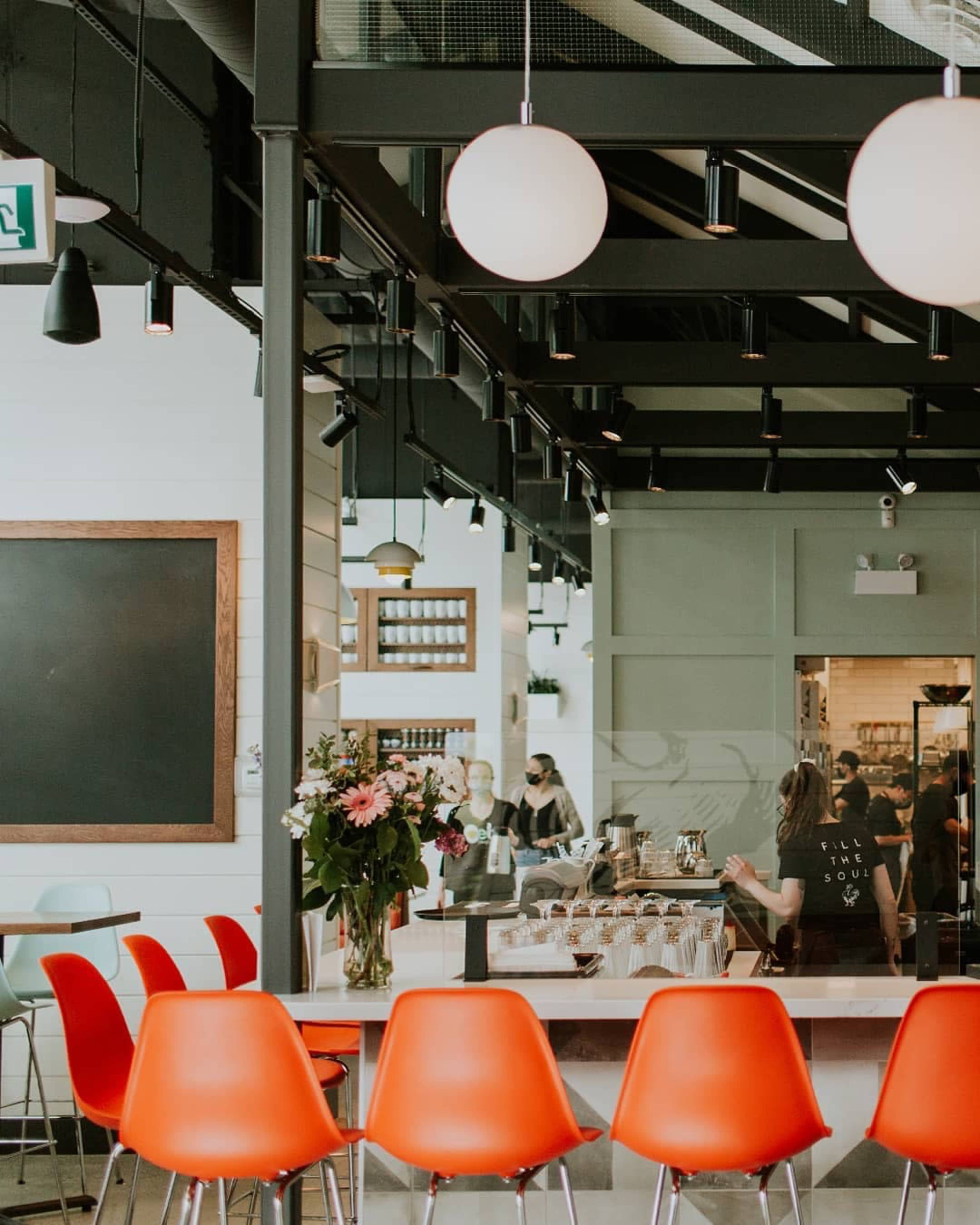 The image shows a modern café interior with a row of orange chairs at a counter, surrounded by various light fixtures and a large chalkboard.