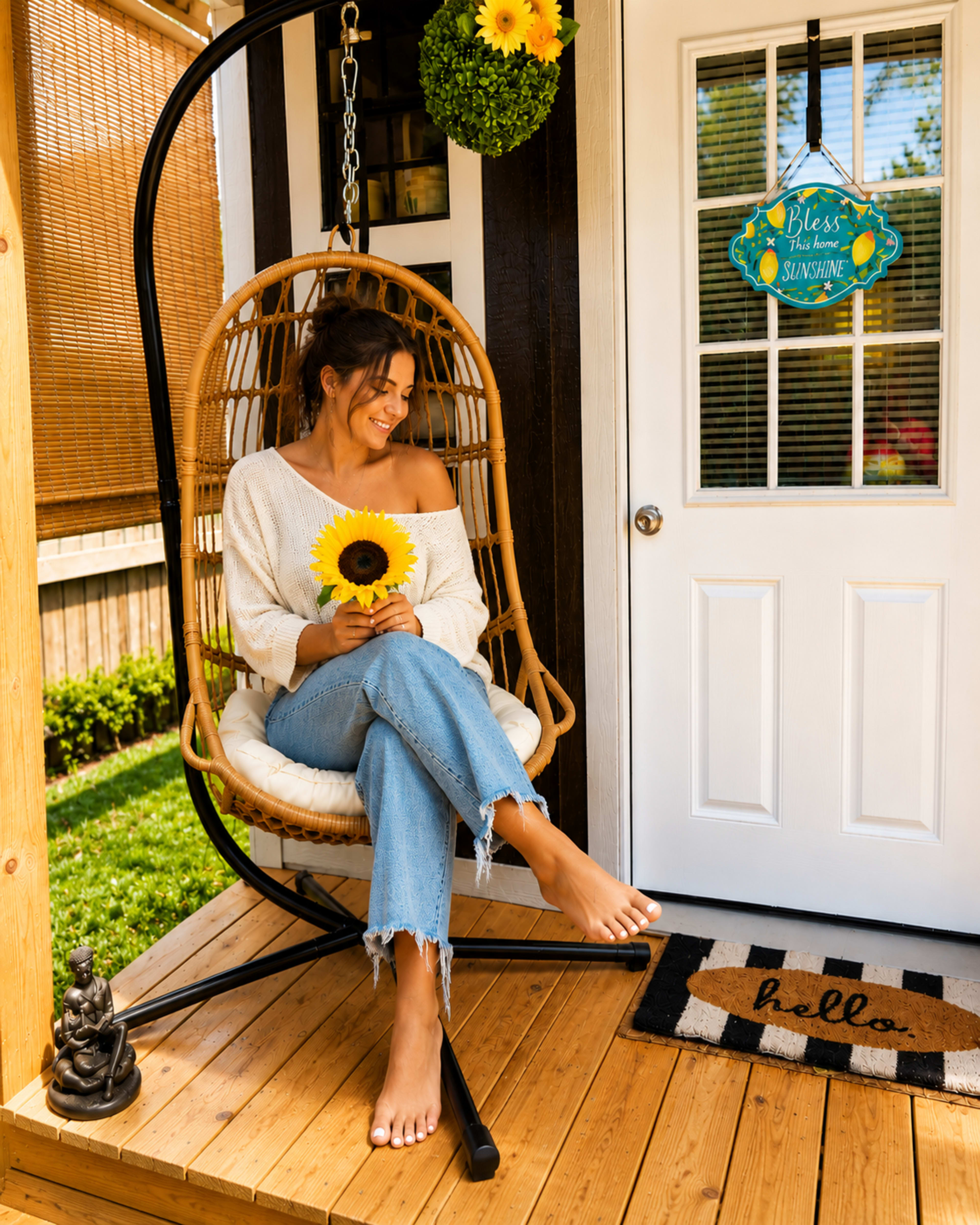 A woman sitting on a hanging chair on a porch holds a sunflower, while a colorful sign hangs on the door and a small statue is nearby.