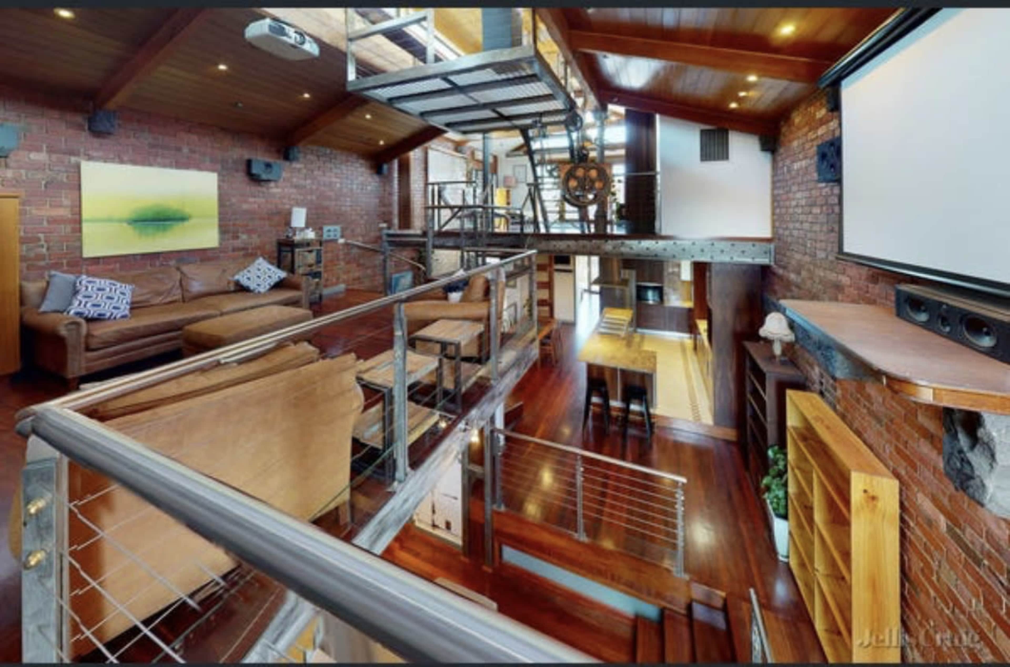 A spacious industrial-style living room with exposed brick walls, wooden floors, and a large staircase leading to an upper level.