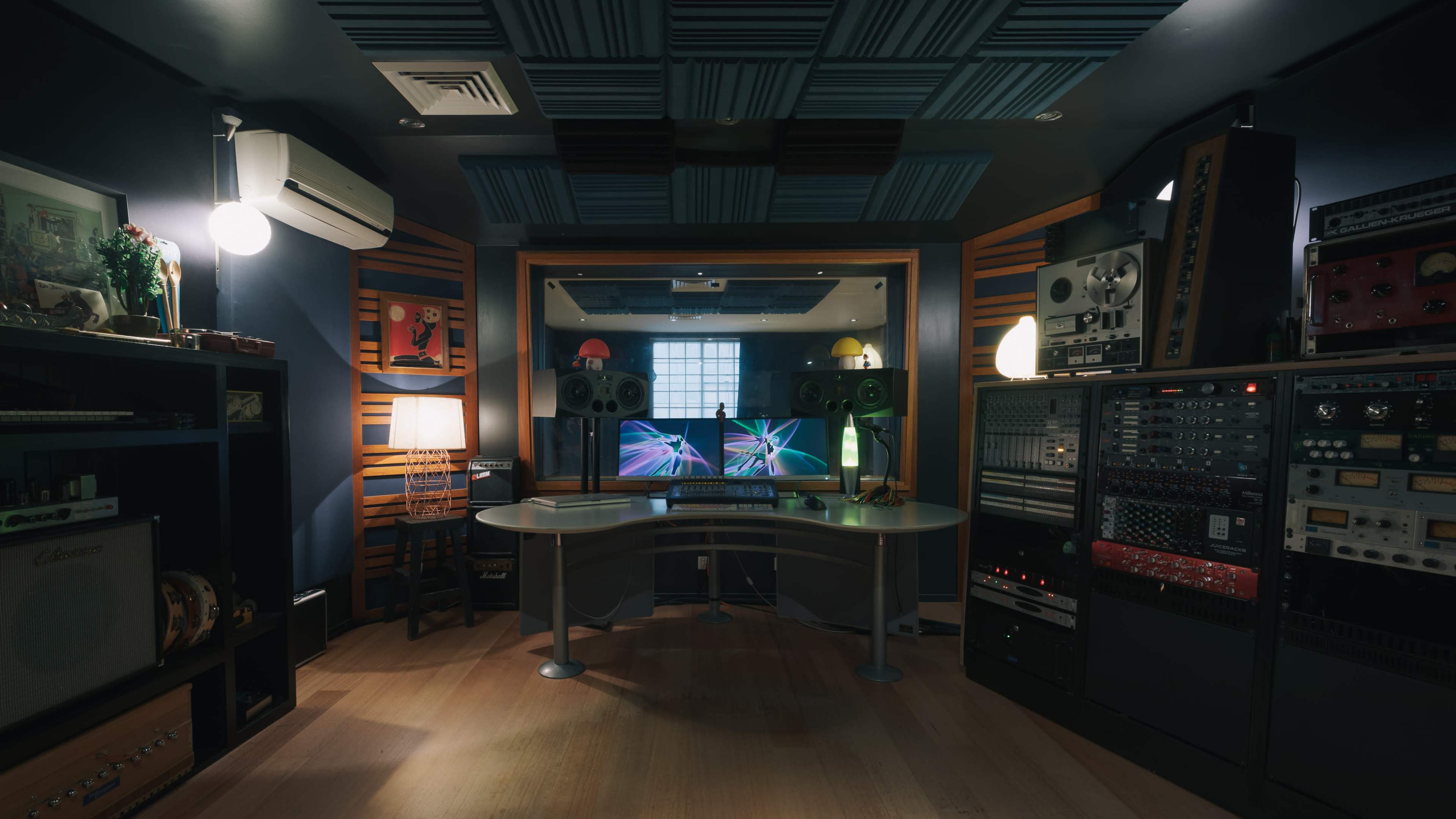A sound recording studio features a central workstation with two monitors, flanked by audio equipment and soundproofing panels on the walls.