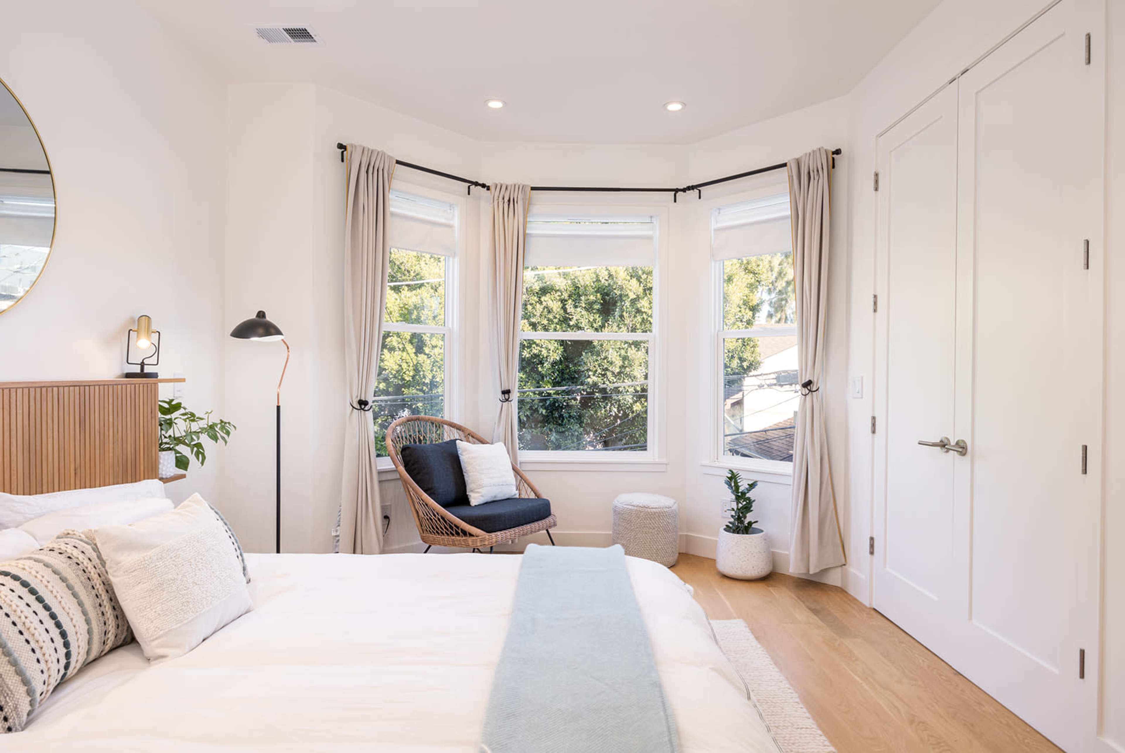 A bright bedroom with a large bed, a cozy reading nook by the windows, and a couple of plants.