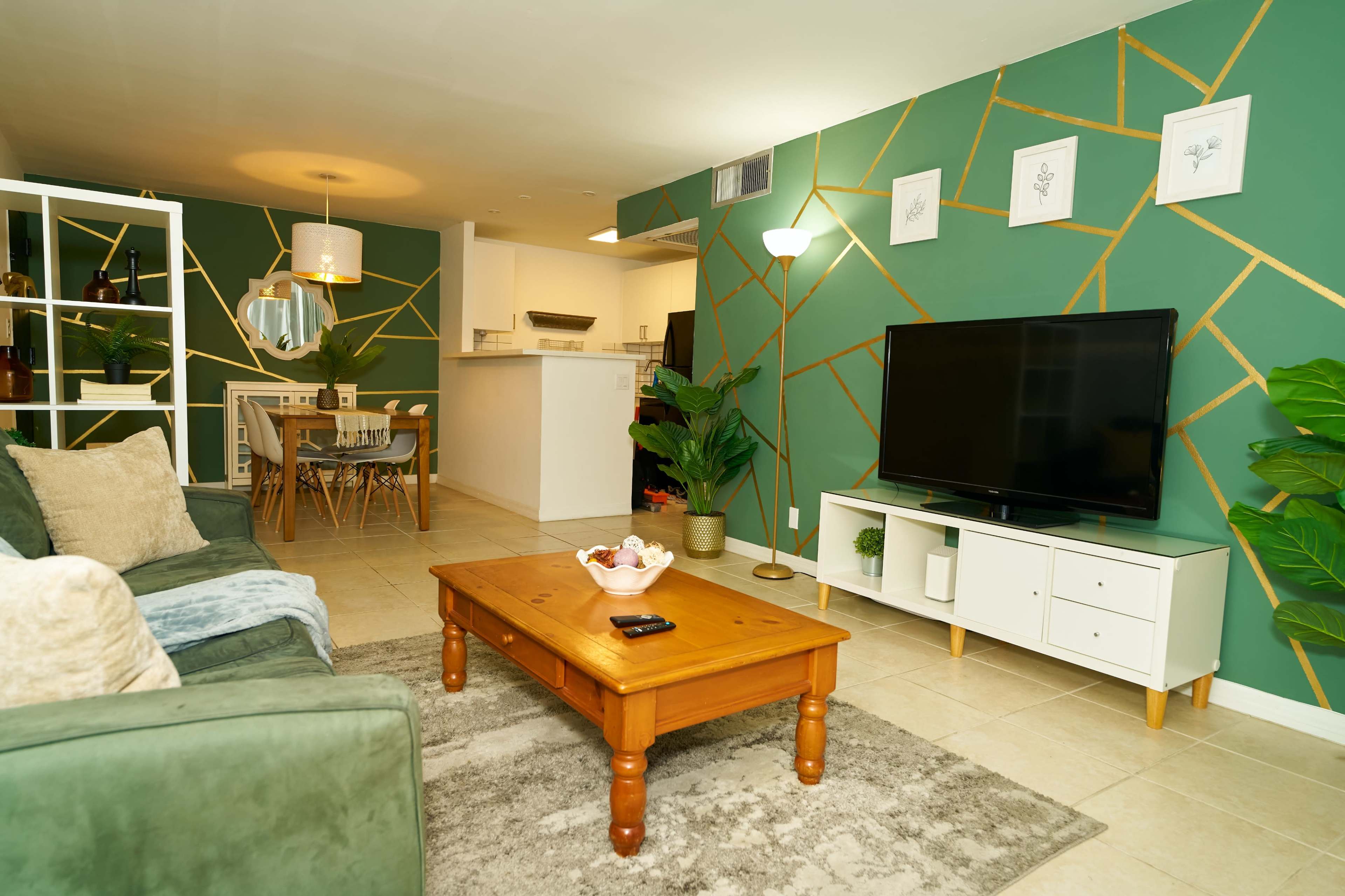 A modern living room features a green accent wall with gold geometric lines, a wooden coffee table, a television on a white stand, and a dining area visible in the background.
