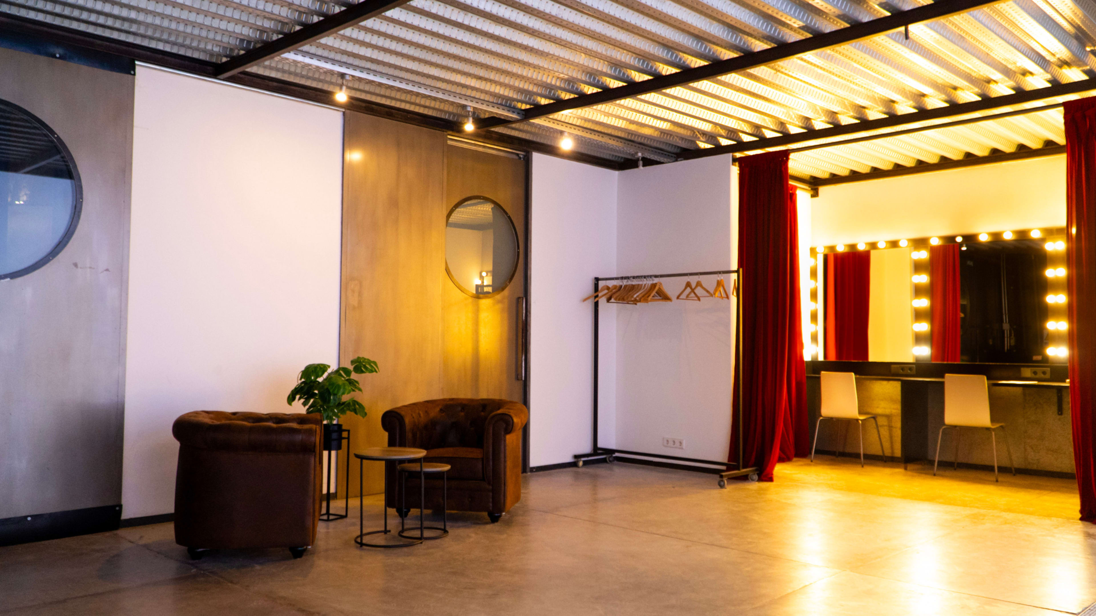 The image shows a spacious room with a seating area, a decorative plant, and a well-lit vanity mirror against the wall.