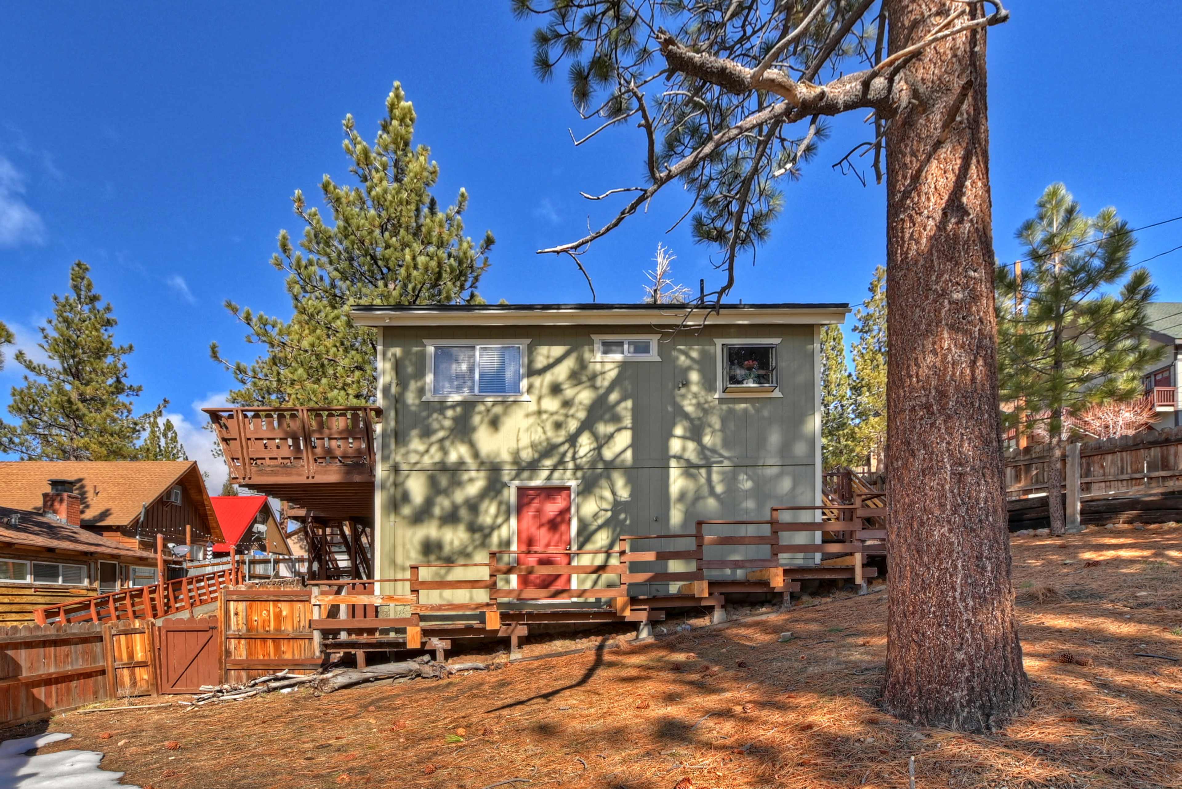 A two-story green house with a red door and wooden staircase is situated on a hillside surrounded by pine trees.