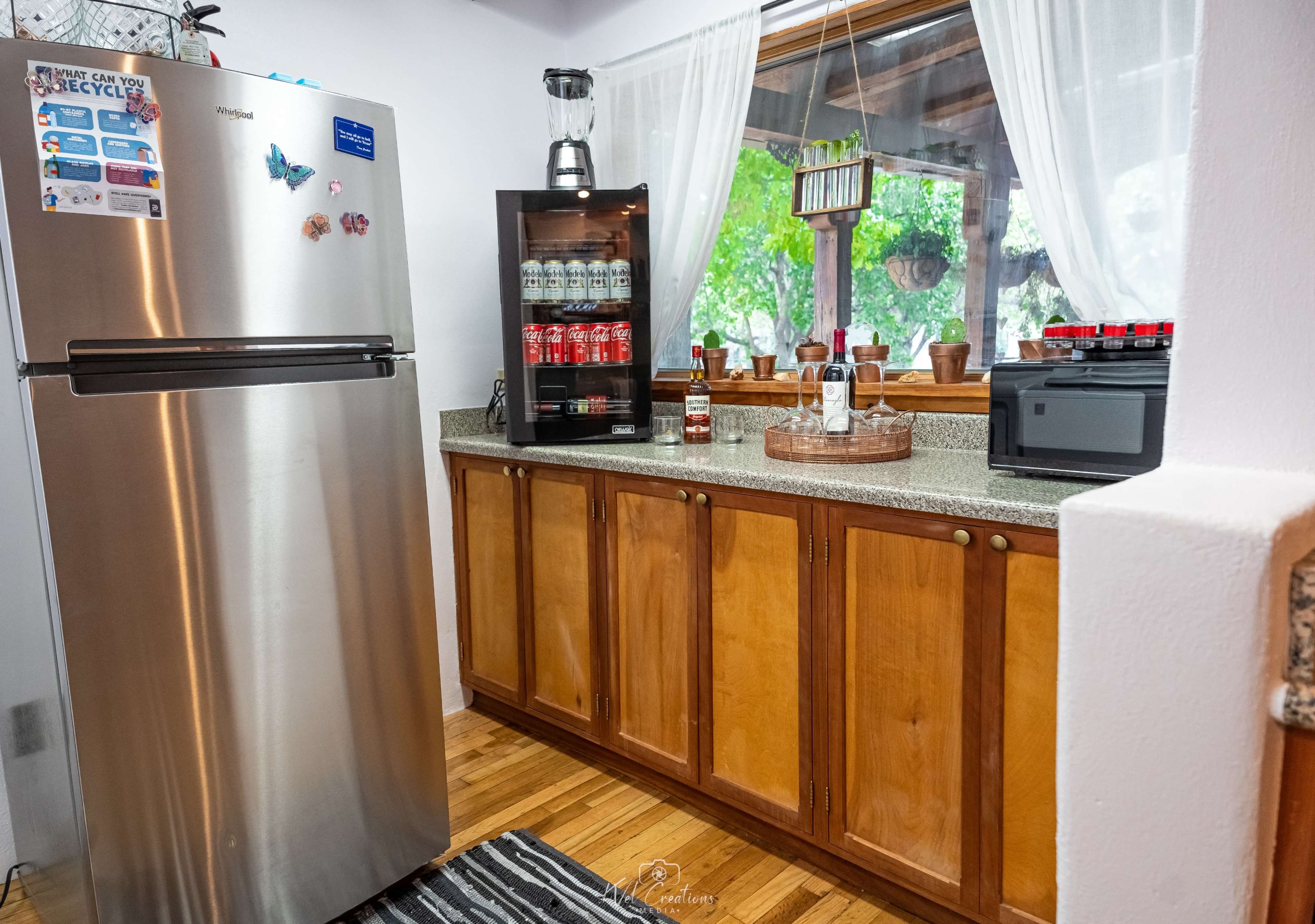 The image shows a kitchen corner with a stainless steel refrigerator, a countertop with a variety of drinks in a glass cabinet, and a window with sheer curtains.
