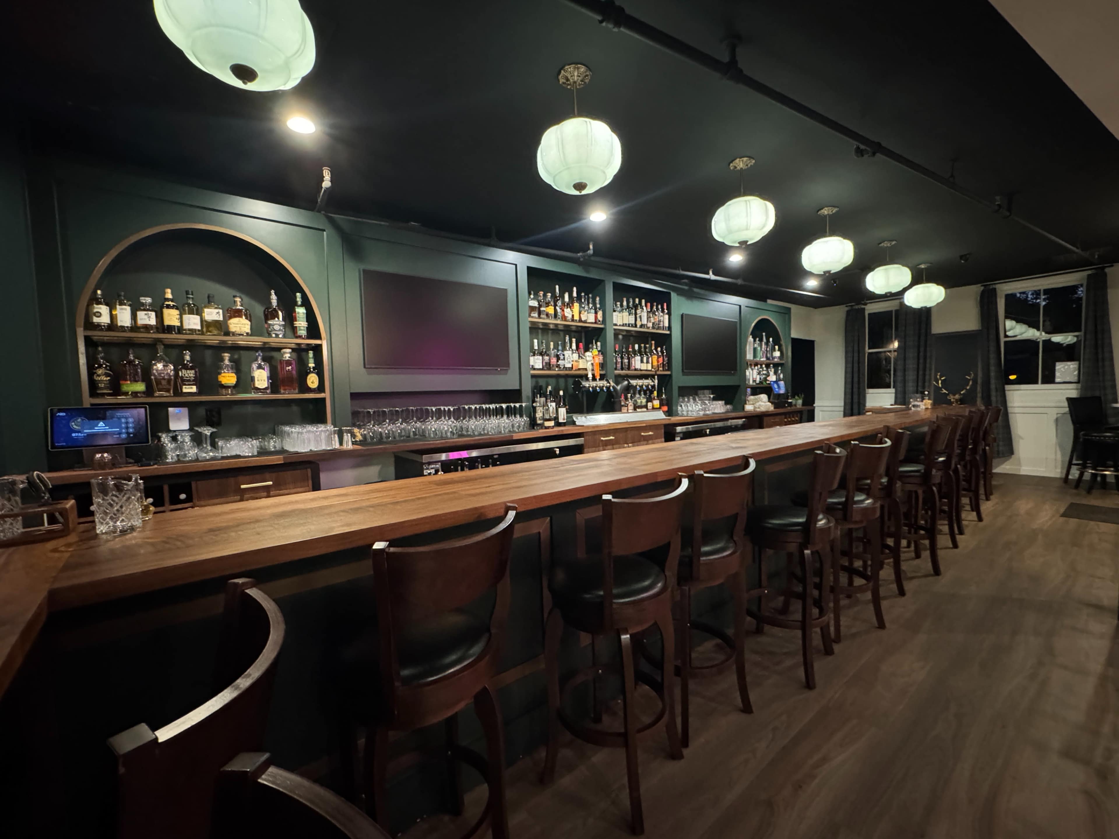 The image shows a stylish bar area featuring a long wooden counter, shelves stocked with various bottles, and hanging green lantern lights.