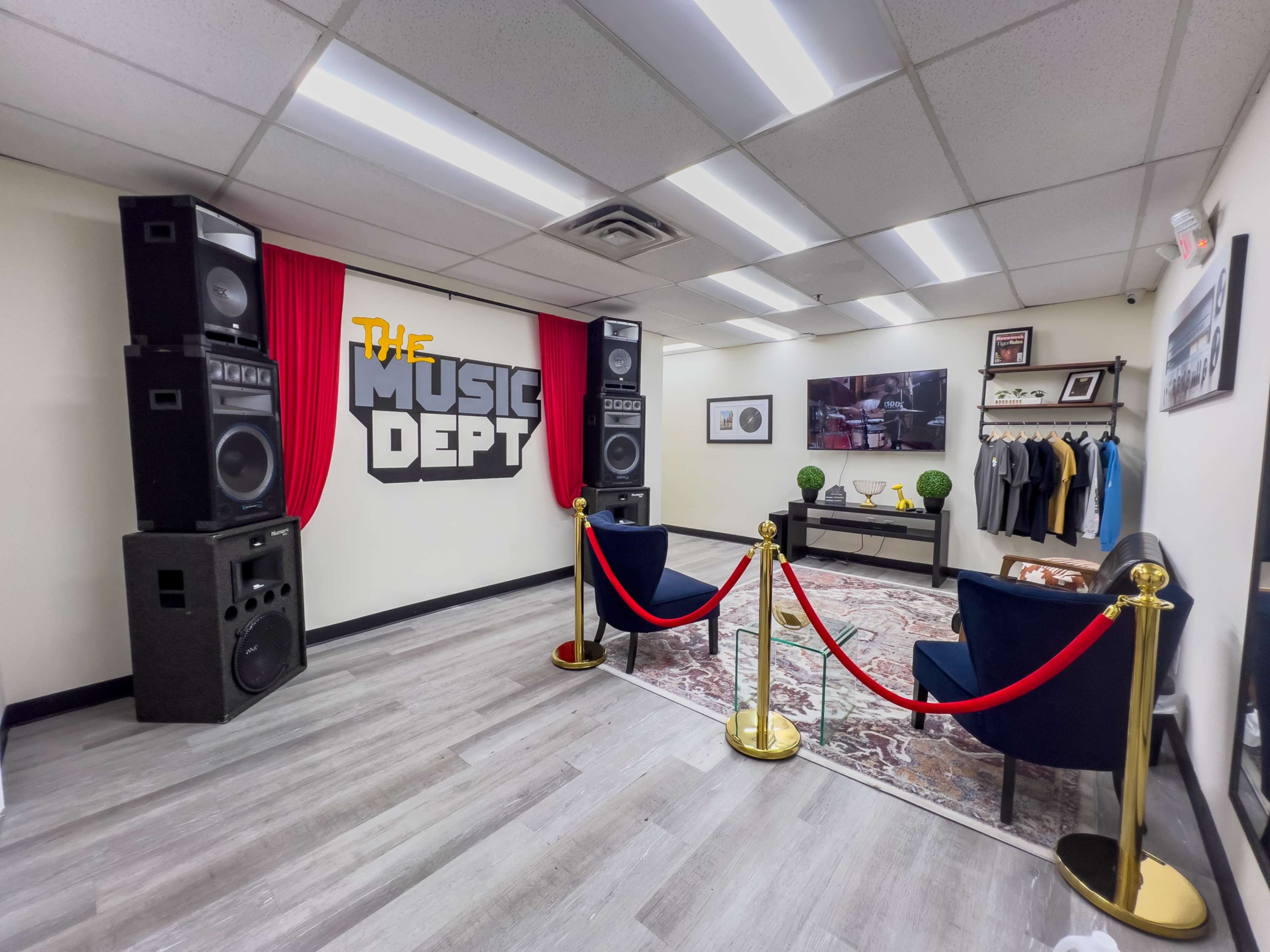 A modern room featuring large speakers on either side of a red curtain, a decorative rug, two blue chairs, a clothing rack, and a wall with "THE MUSIC DEPT" displayed prominently.