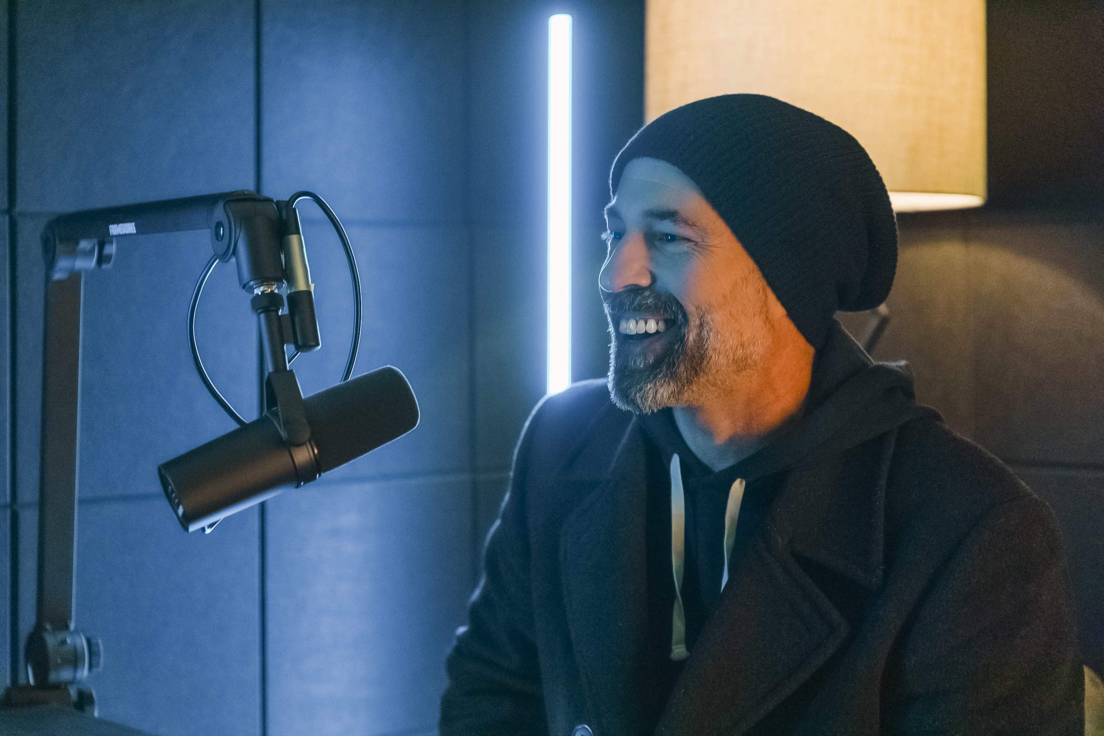 A man with a beard and beanie sits in front of a microphone in a dimly lit room.