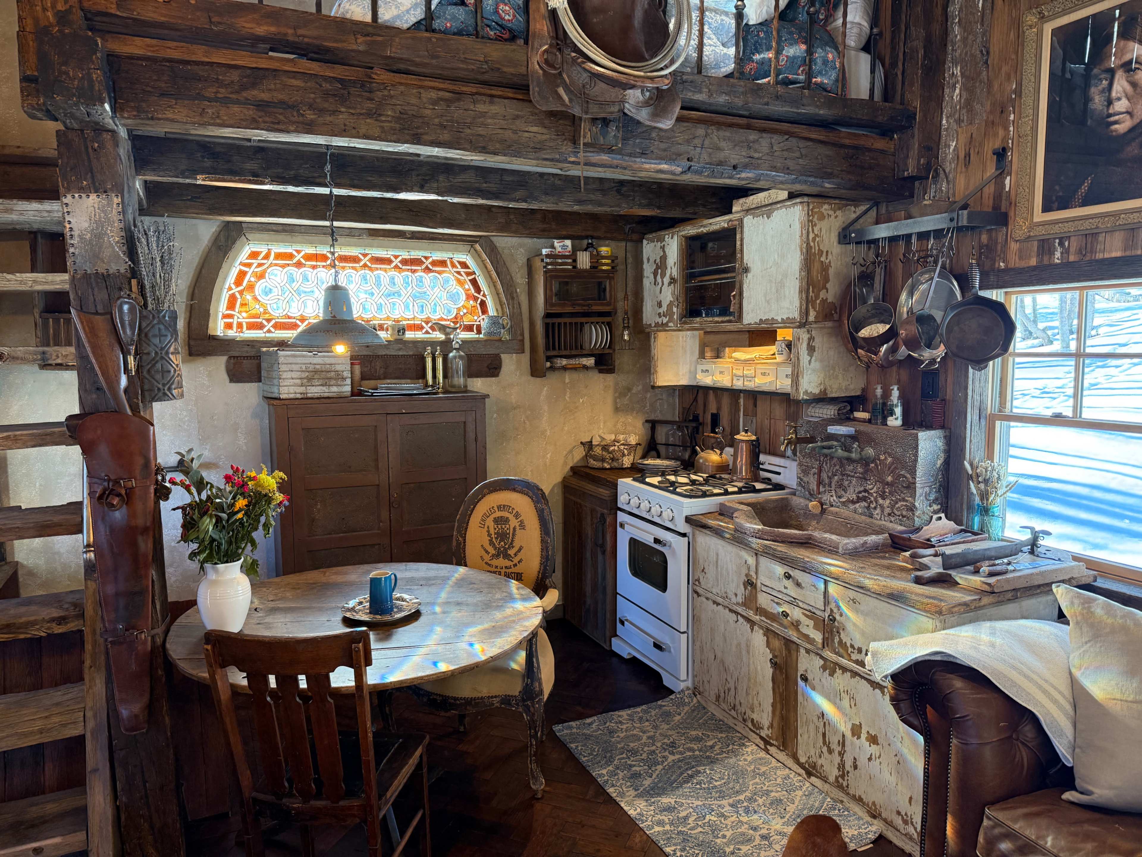 The image shows a rustic kitchen with a wooden table, vintage appliances, and a staircase leading to a loft area.