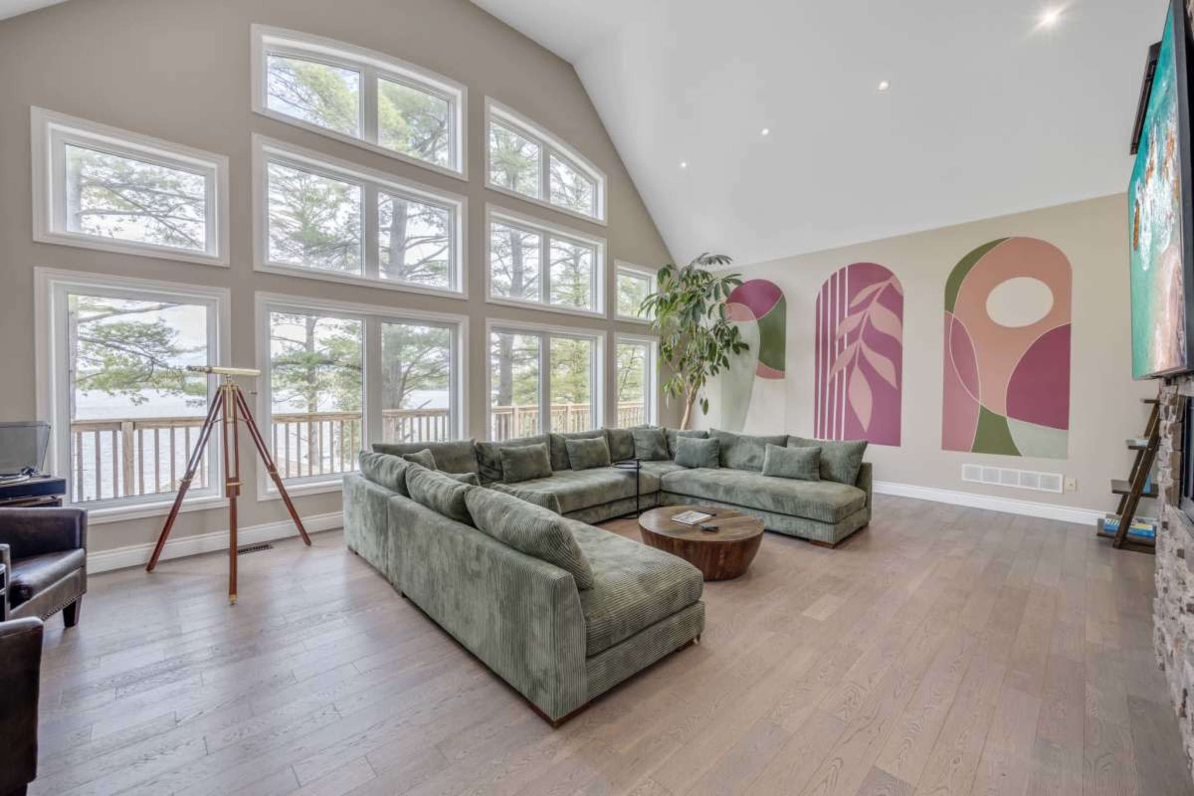 A spacious living room with large windows overlooking a lake, featuring a green sectional sofa, a wooden coffee table, and abstract wall art.