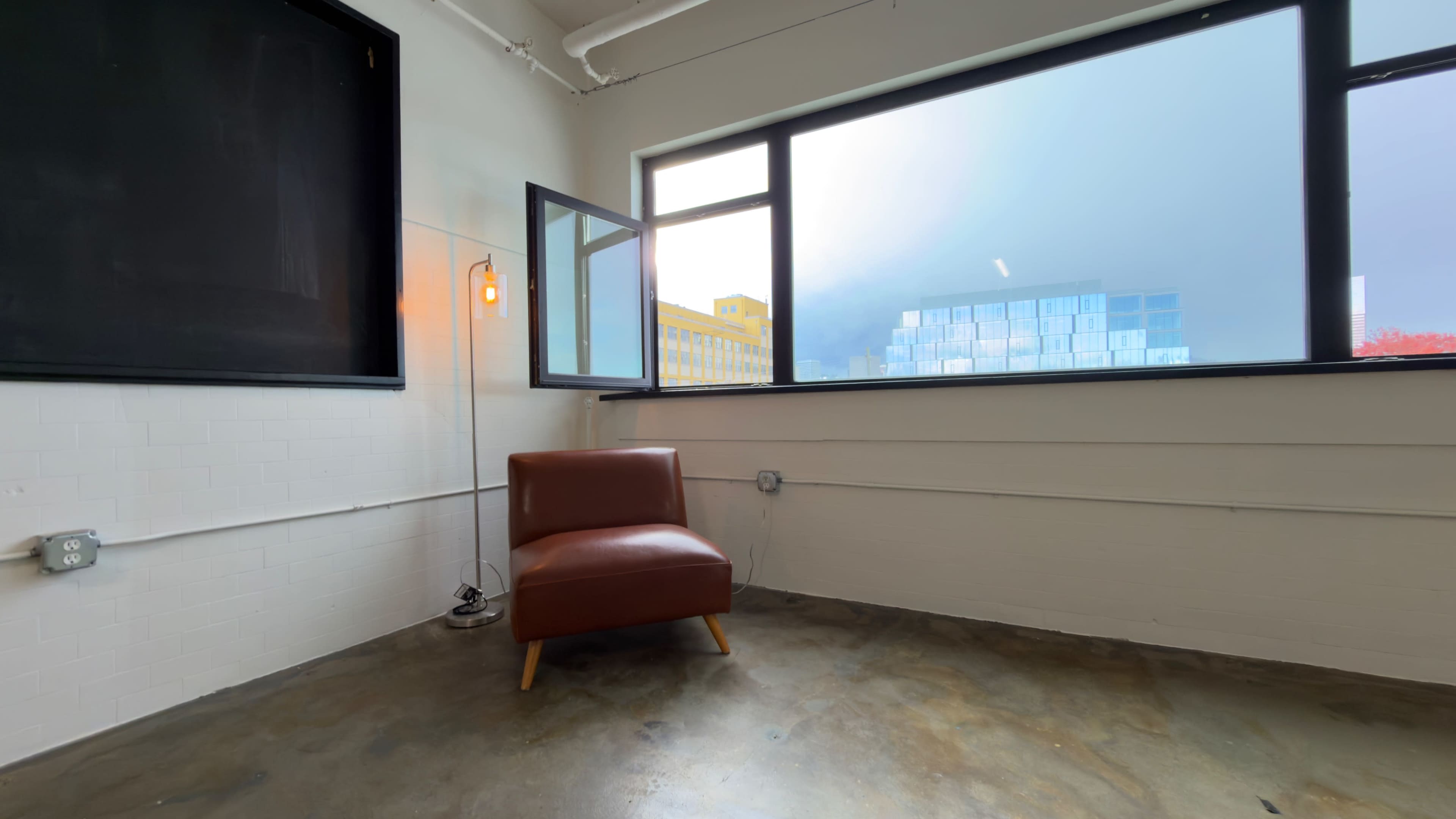 A brown armchair sits in the corner of a room with large windows, showing a view of a building outside.