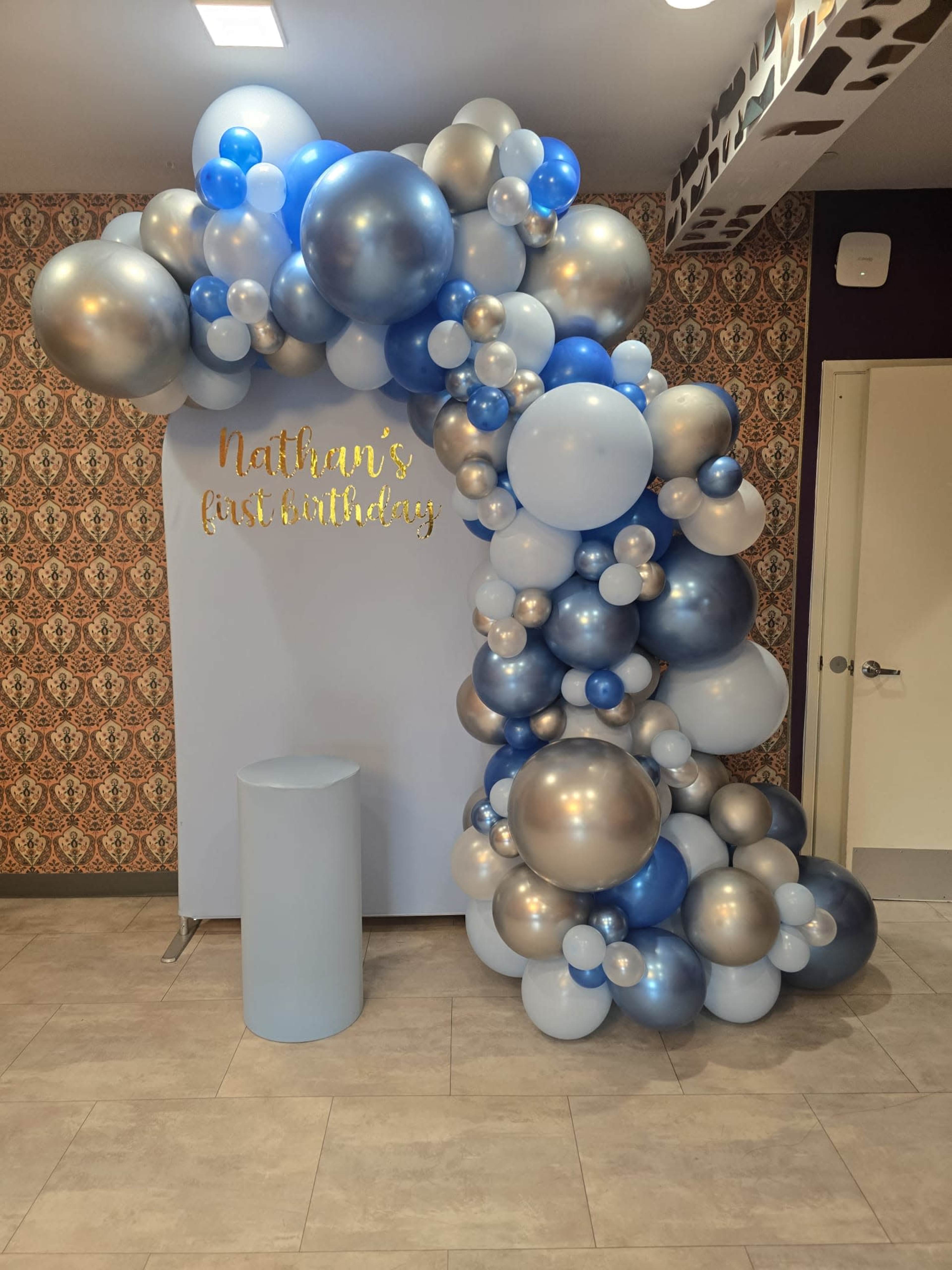 The image shows a decorative balloon arch in shades of blue and silver, set against a wall with gold lettering that reads "Nathan's first birthday."