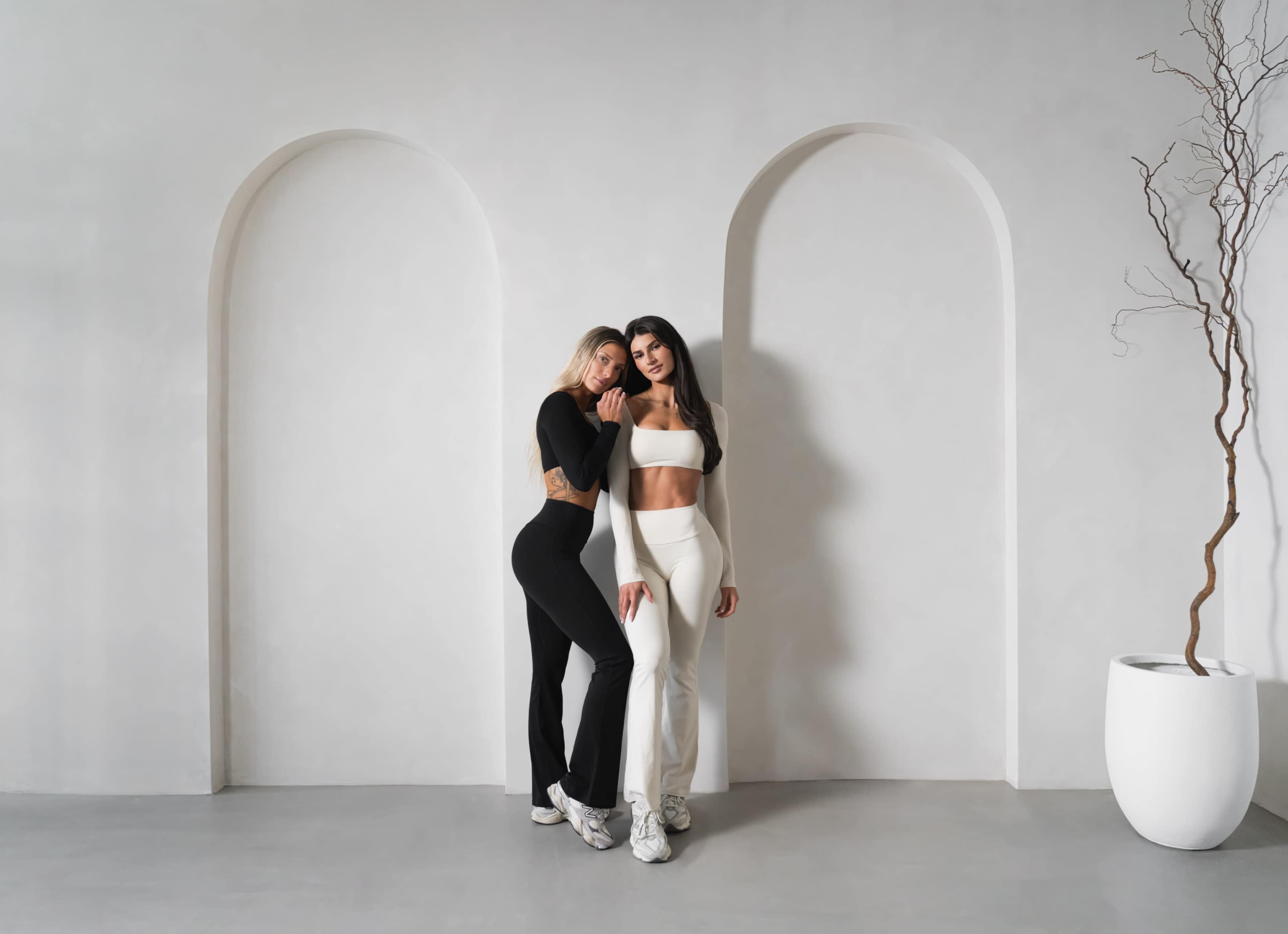 Two women pose in fitted athletic outfits against a minimalist backdrop with arched openings and a potted plant.