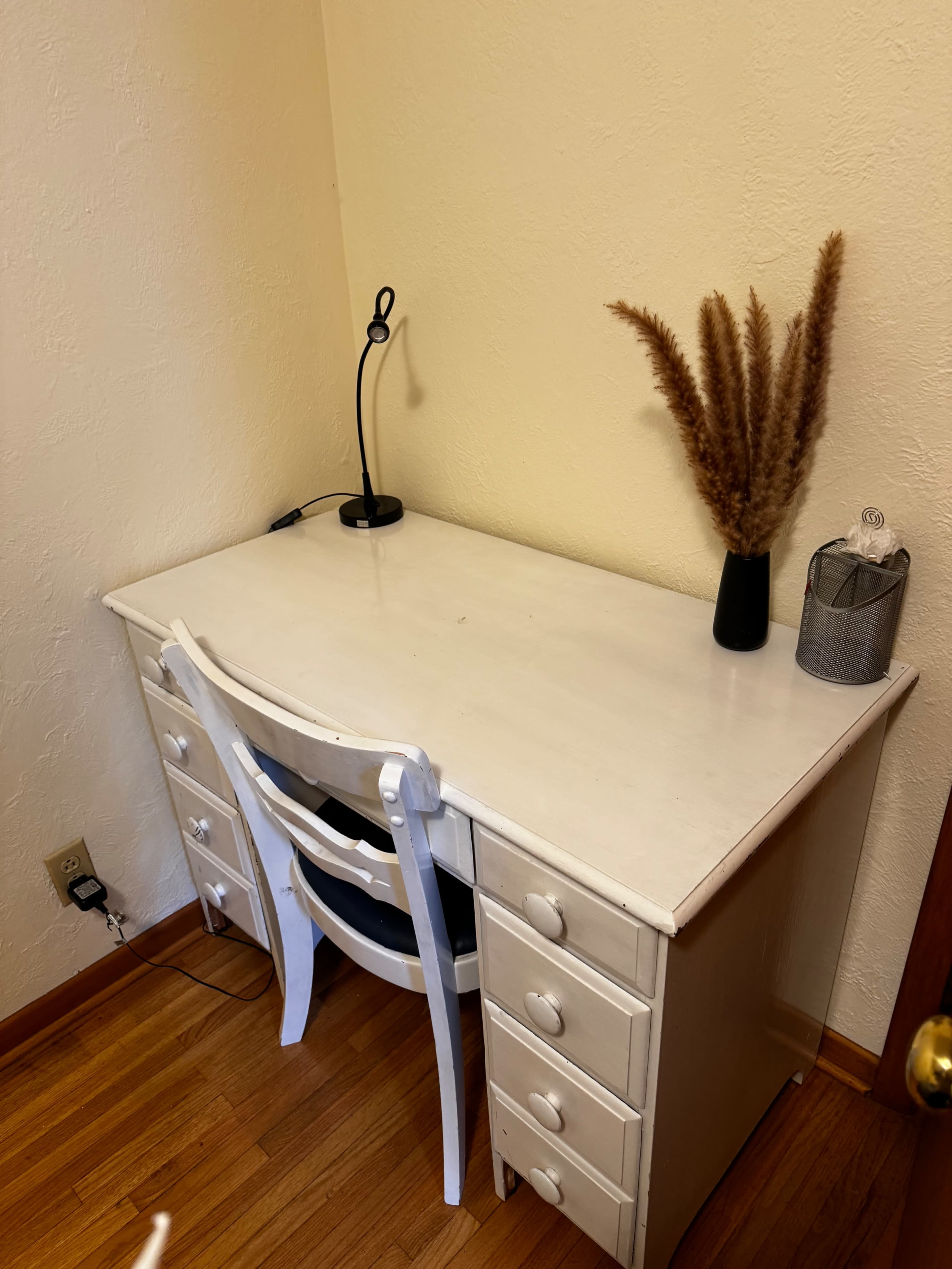 A white desk is positioned against a beige wall, featuring a chair, a table lamp, and a vase with dried foliage.