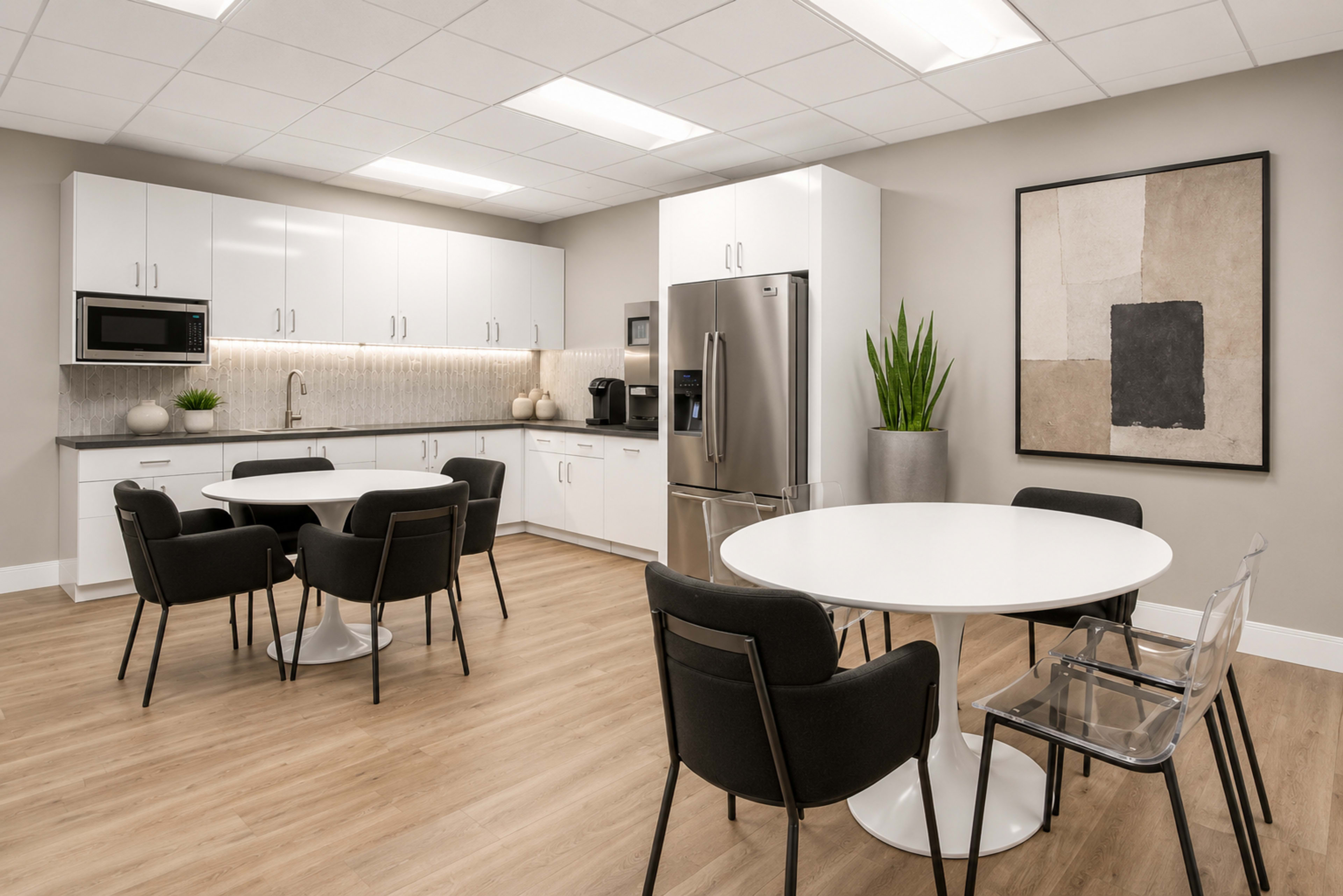 The image shows a modern kitchen and dining area featuring white cabinetry, a stainless steel refrigerator, two circular tables with black chairs, and a large piece of wall art.