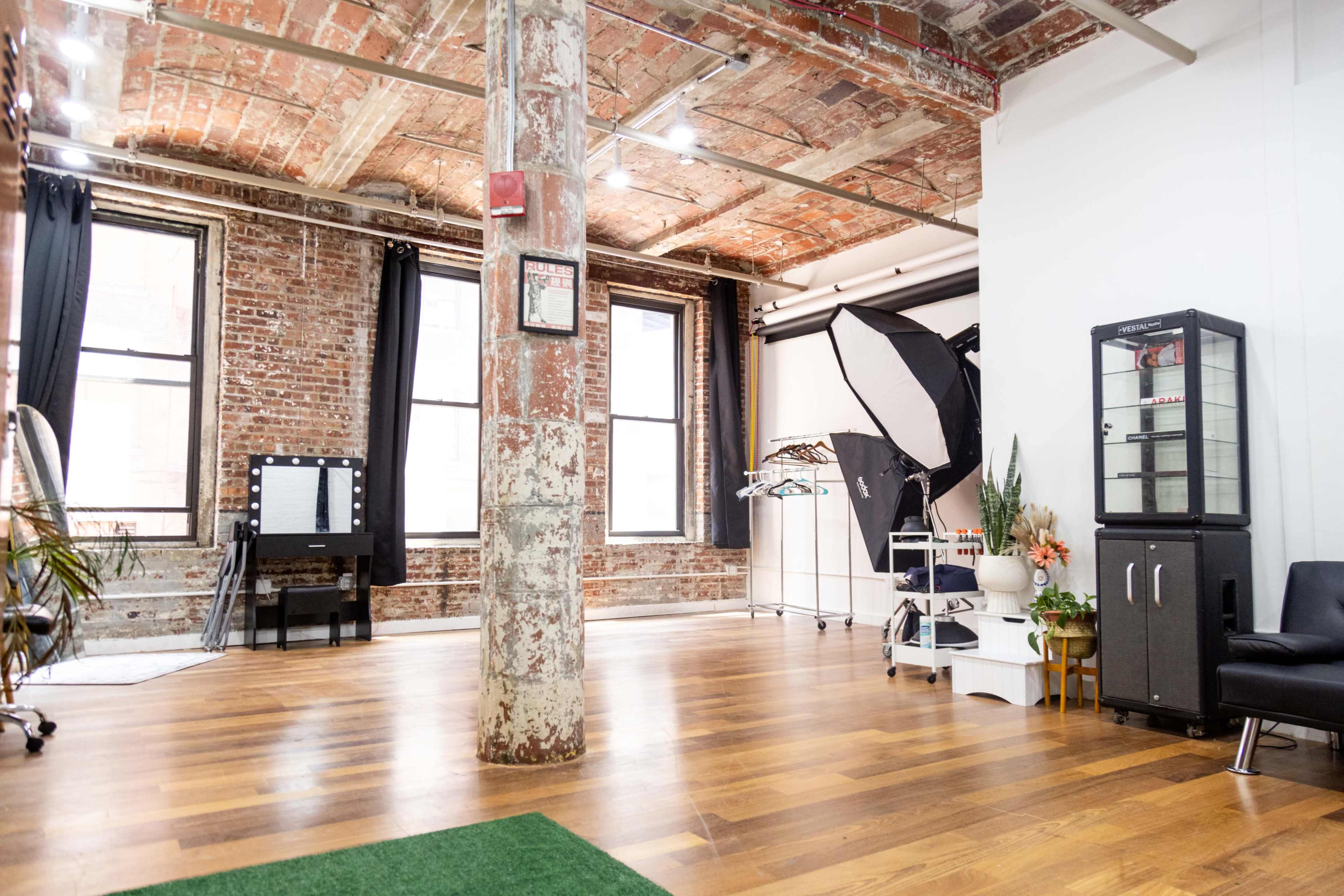 An industrial-style room with exposed brick walls, wooden flooring, and various photography equipment, including a reflector and a rack of clothes.