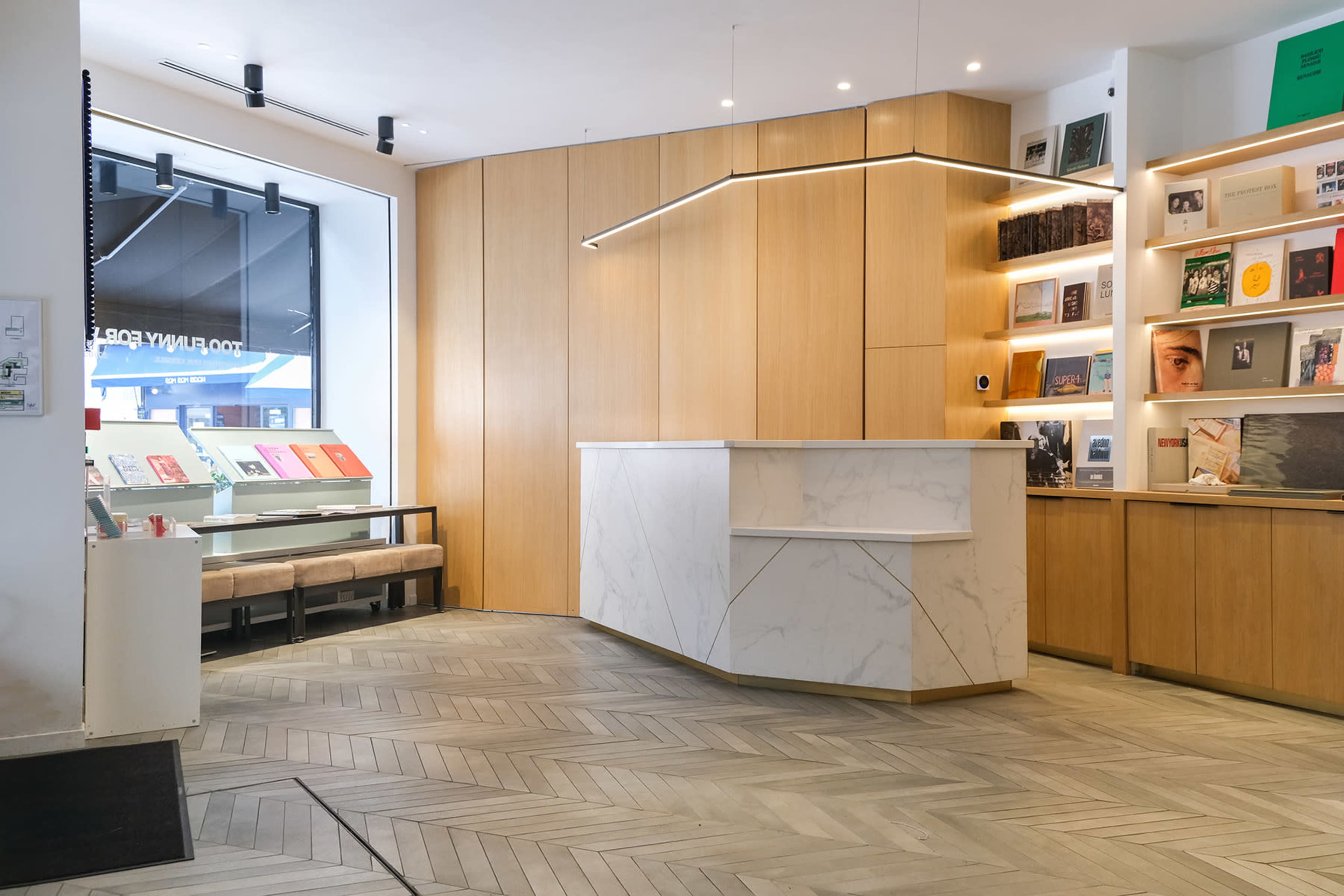 The image shows a modern reception area featuring a marble front desk, wooden paneling, and a display shelf lined with books and magazines.