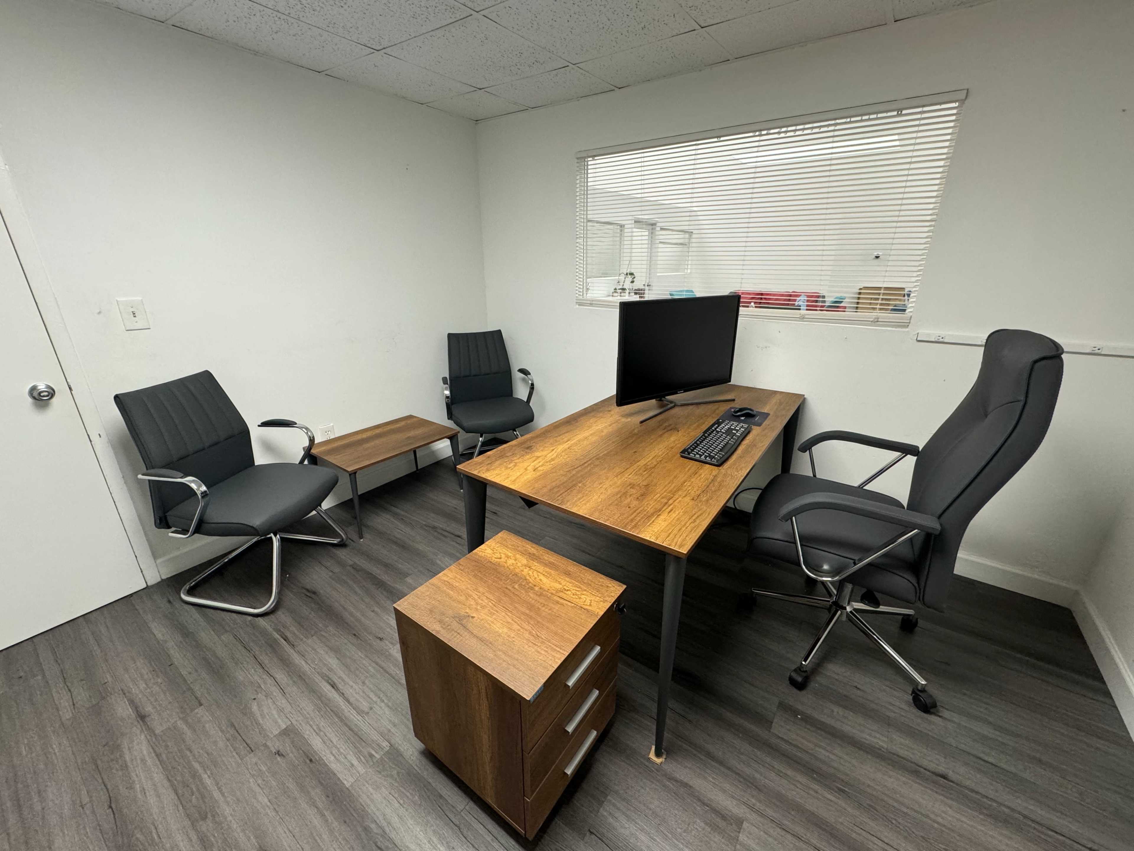 An office with a wooden desk, a computer monitor, and four chairs arranged around it, with a small storage unit nearby.