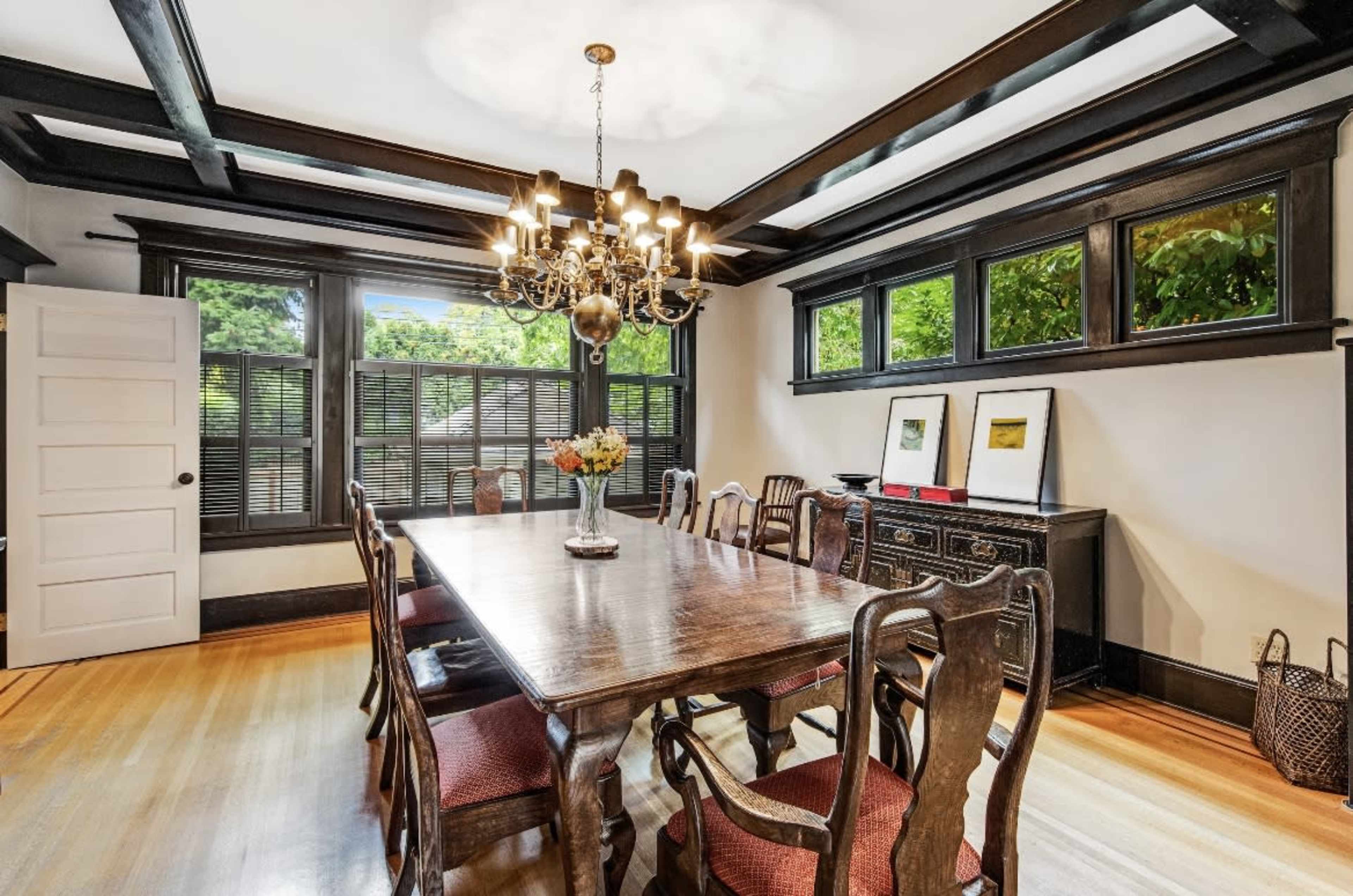 A wooden dining table with several chairs is set in a well-lit room featuring a chandelier and large windows.