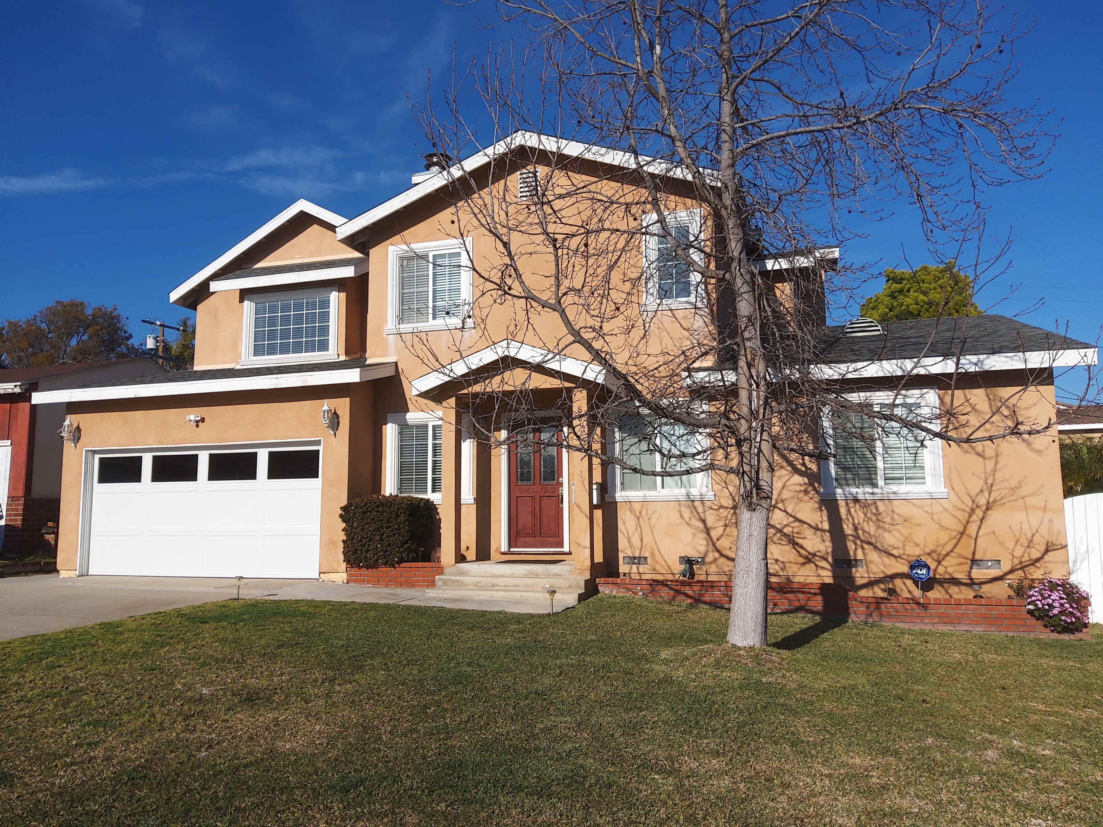 A two-story, tan-colored house with a front porch, a large garage, and a tree in the yard.