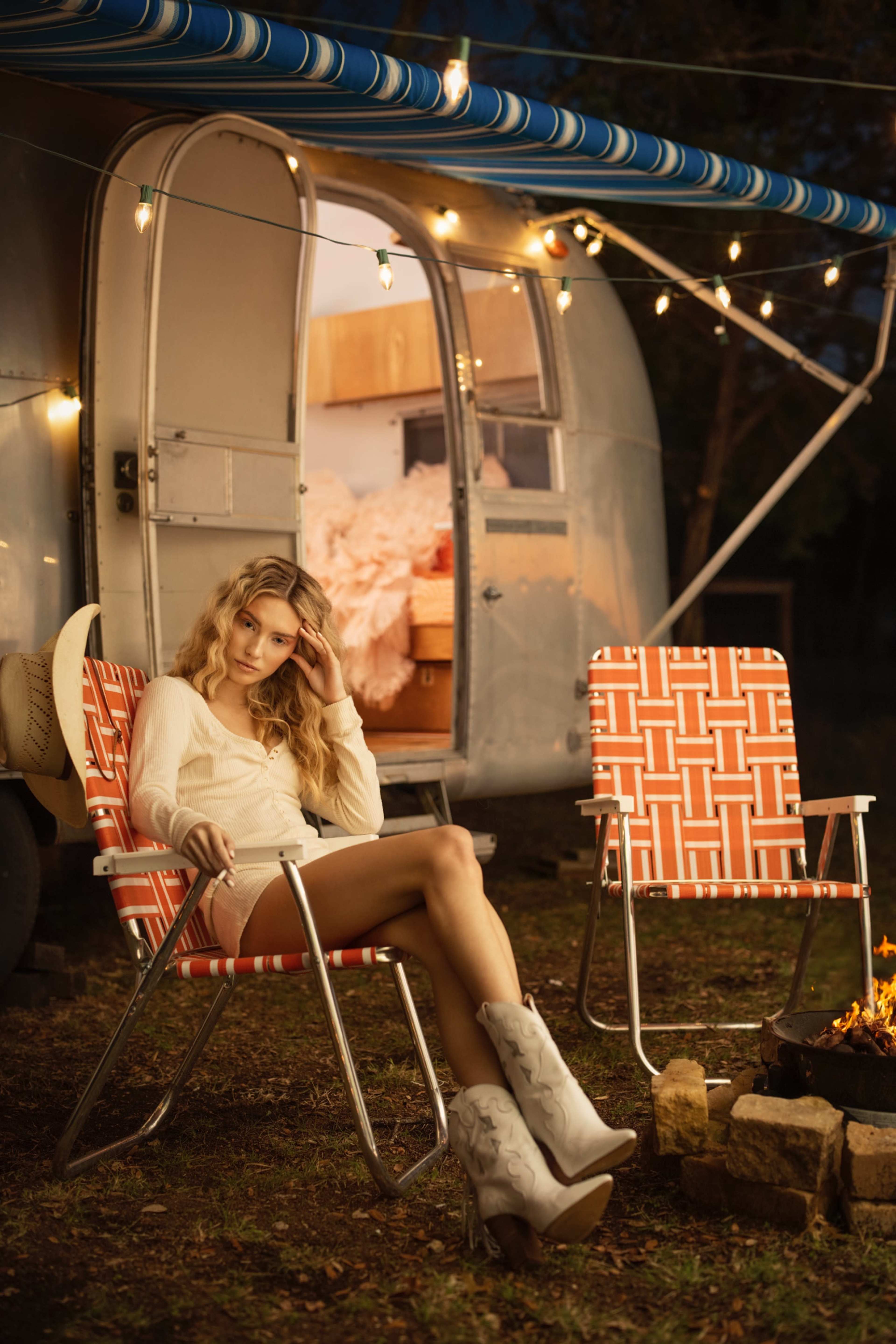 A woman sits on a red and white patterned chair in front of a vintage camper, with a campfire nearby and string lights illuminating the scene.