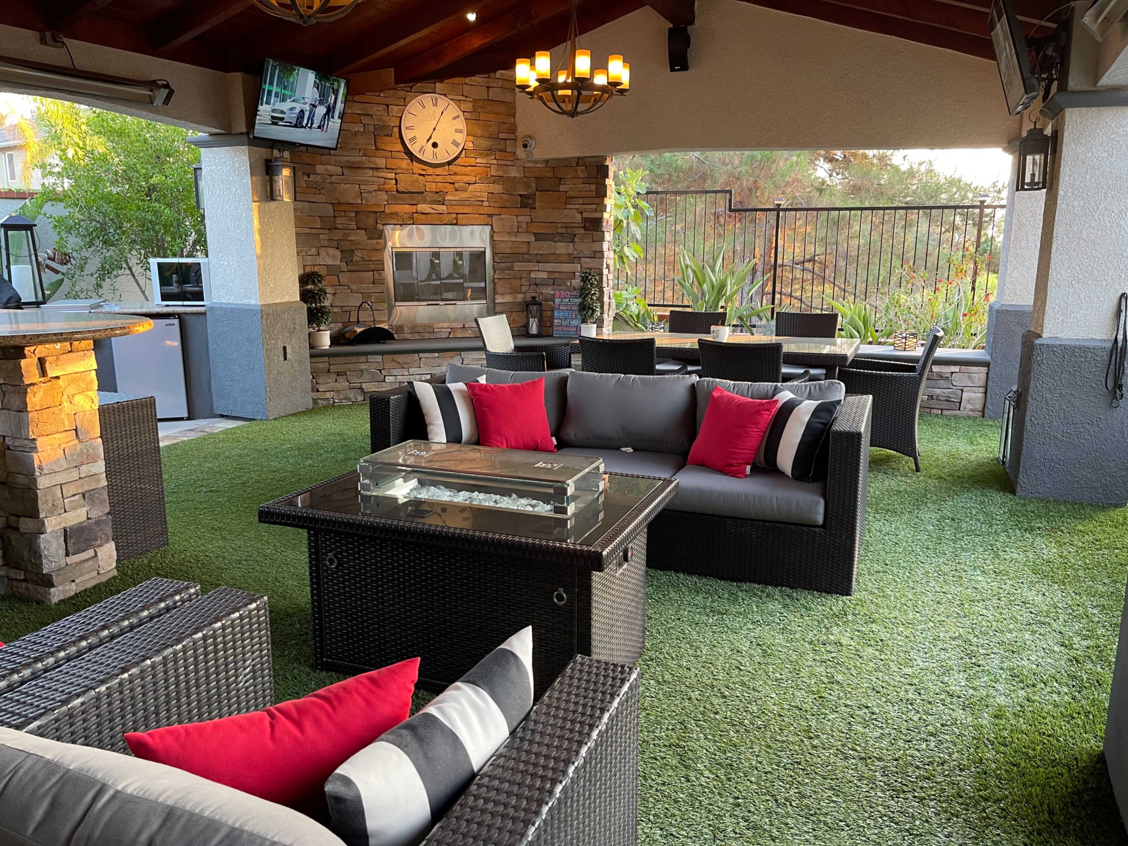 The image shows a patio area furnished with a seating arrangement of dark wicker furniture and red accent pillows, along with a barbecue grill and a stone wall backdrop.
