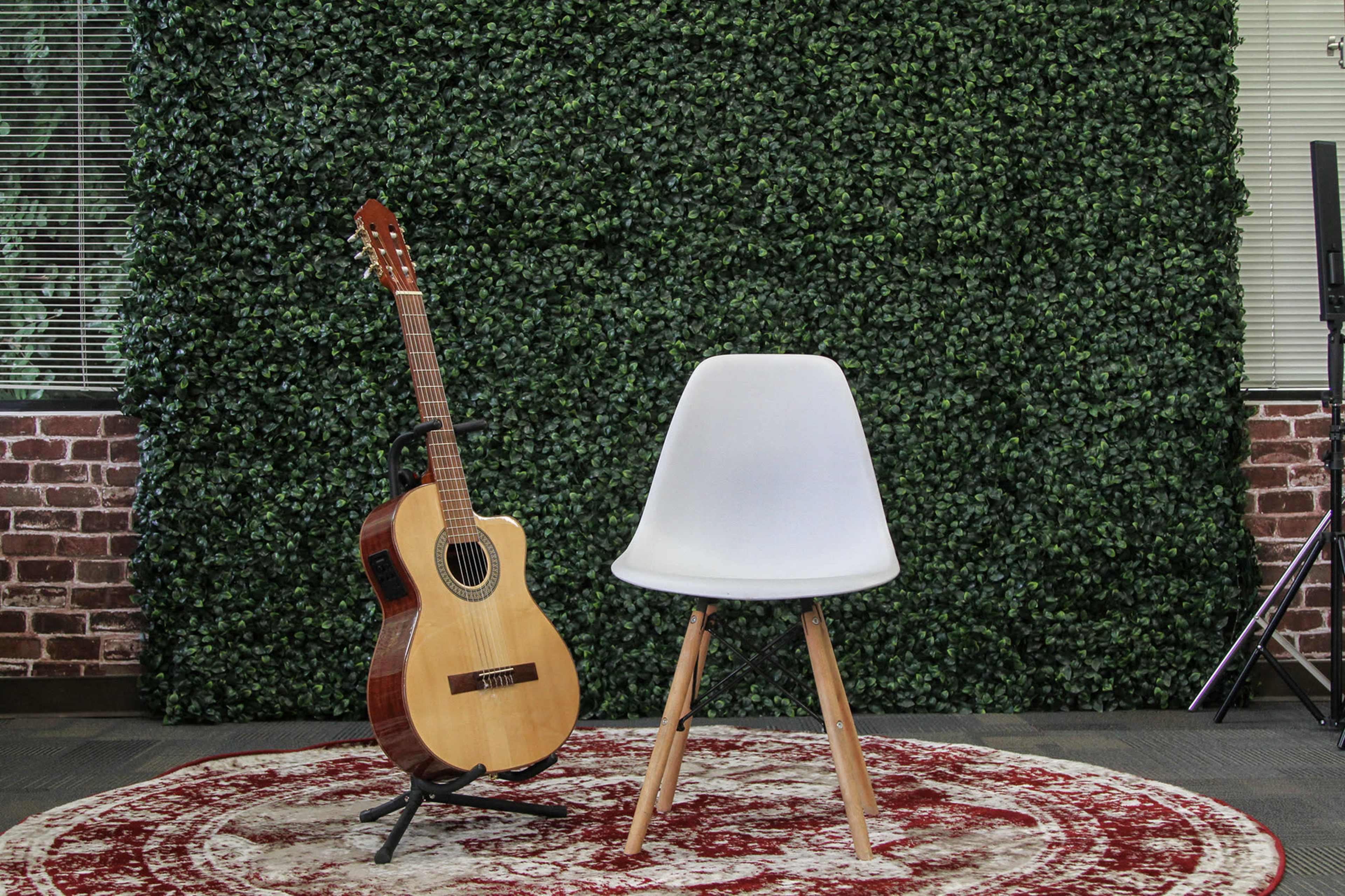 A wooden acoustic guitar leans against a white chair on a patterned rug in front of a green plant wall.