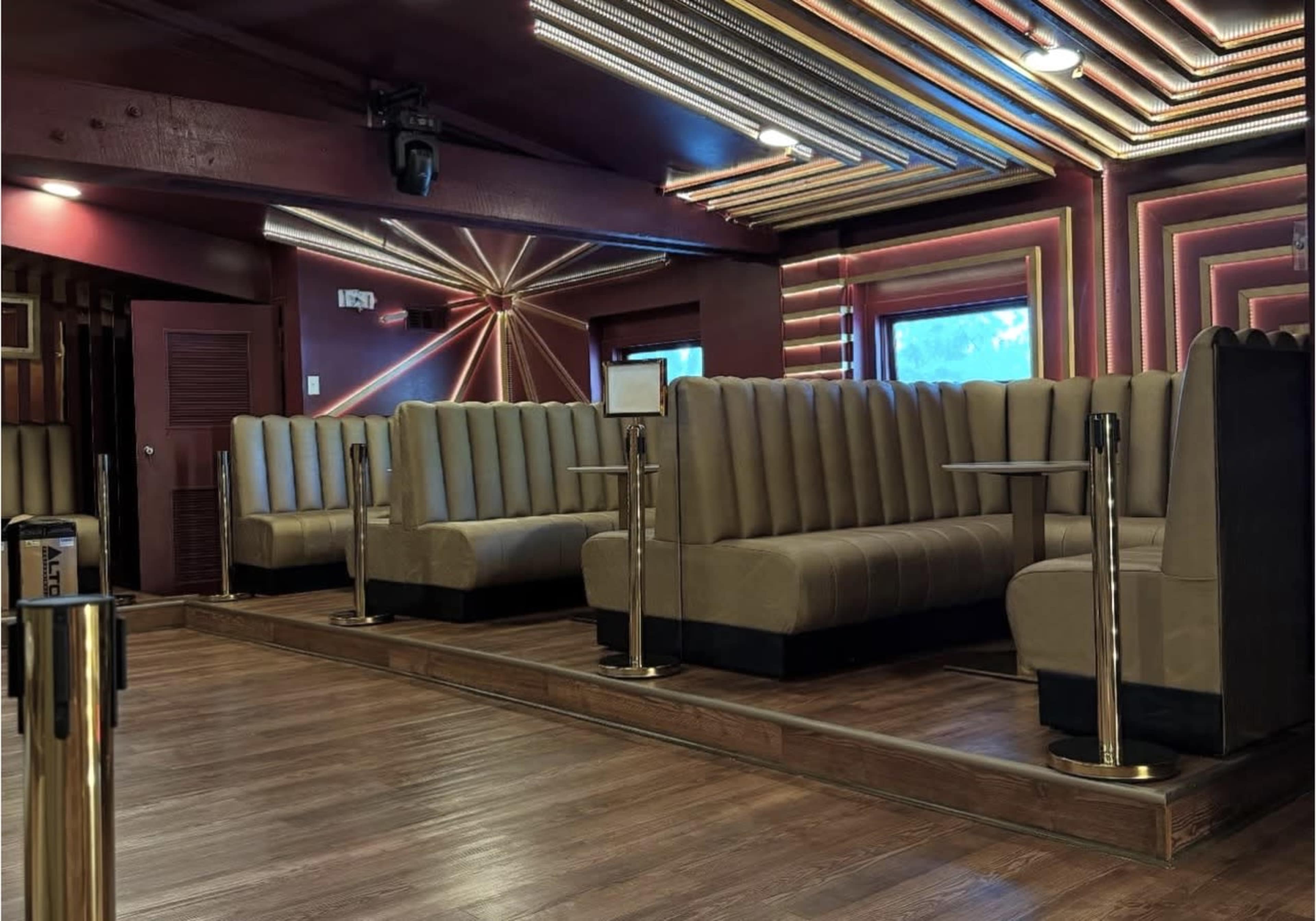 The image shows an interior of a lounge or restaurant featuring plush brown booths with wooden flooring and colorful neon lighting in the ceiling.