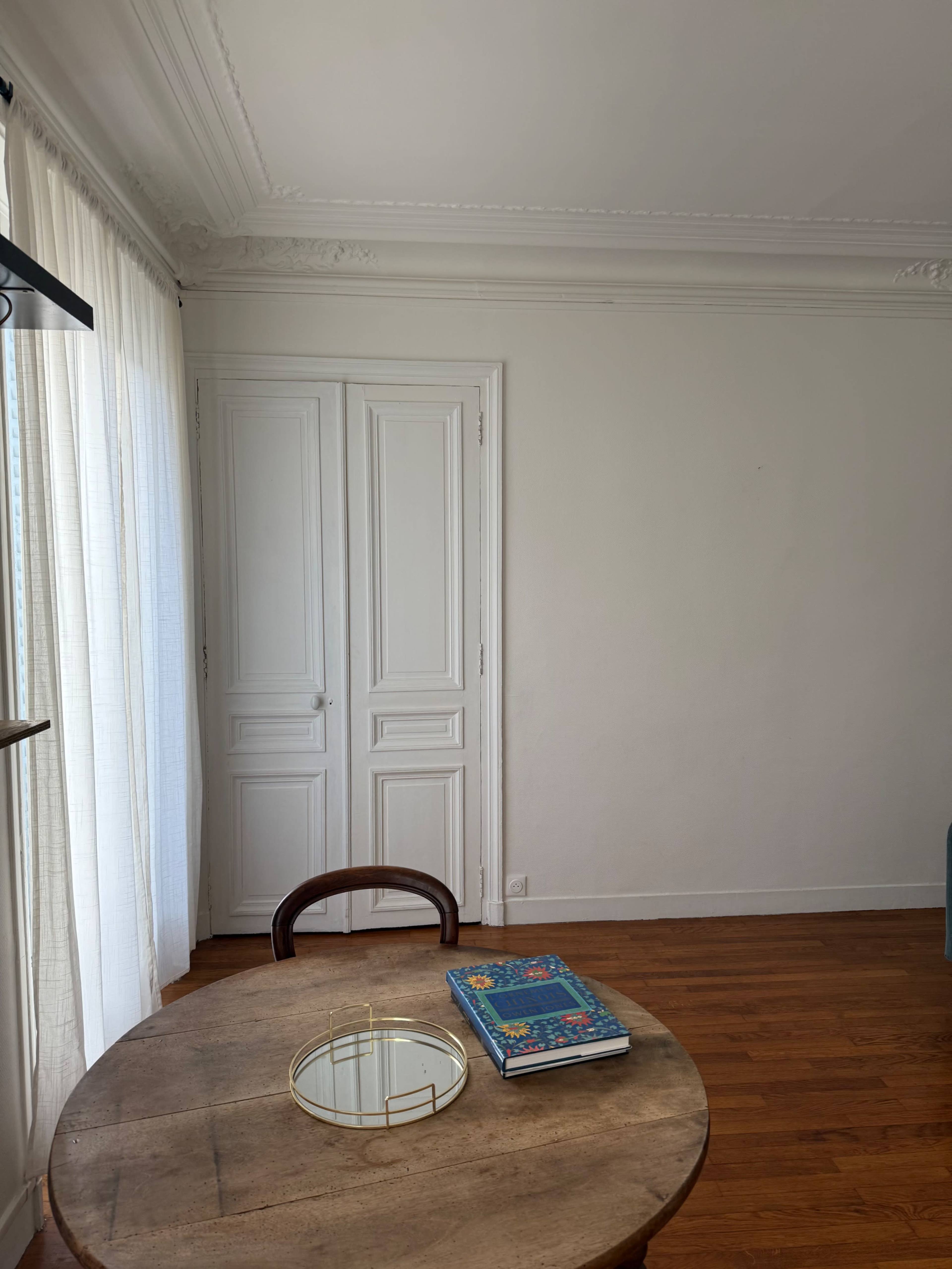 A wooden table with a decorative mirror and a book is set in front of a wall featuring a closed white door, while sheer curtains let in natural light.