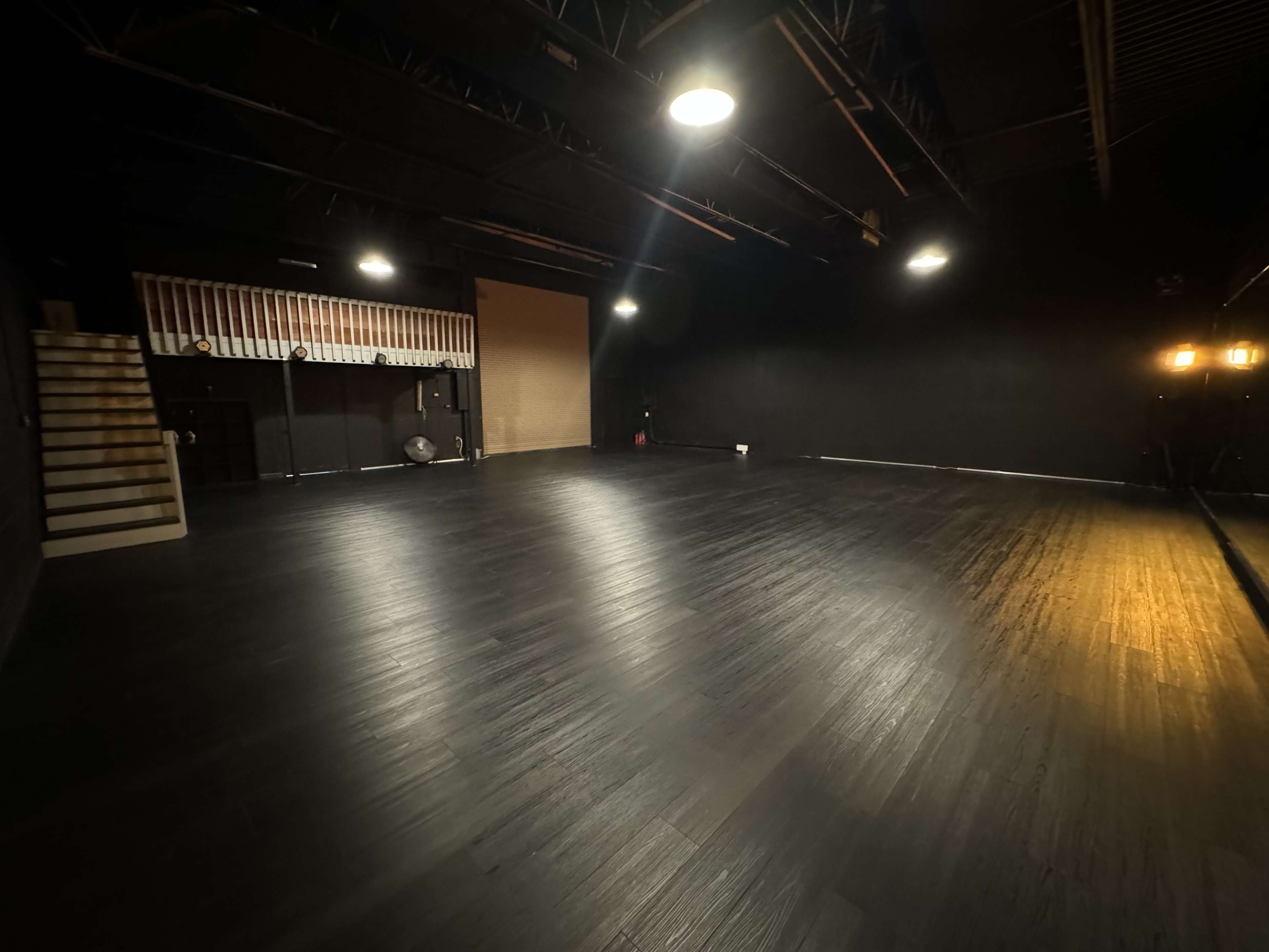 The image shows a spacious, empty black-walled studio with wooden flooring and overhead lights.