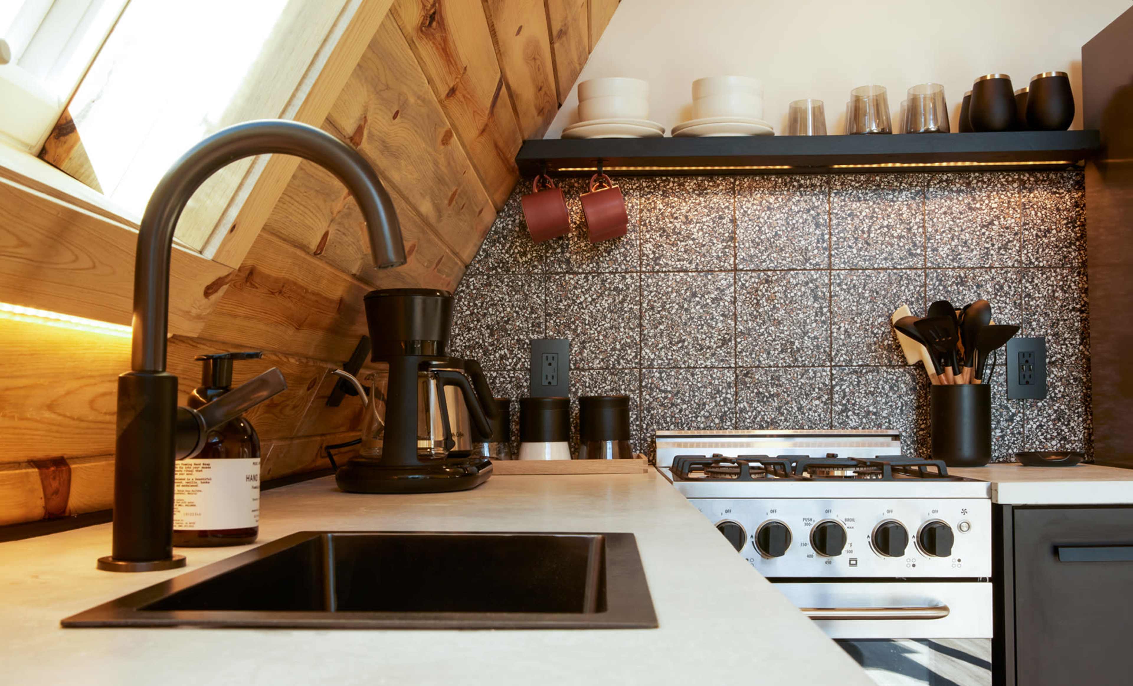 A modern kitchen features a black sink and faucet, a stovetop beneath a backsplash of sparkly tiles, and various kitchen tools neatly arranged on a countertop.