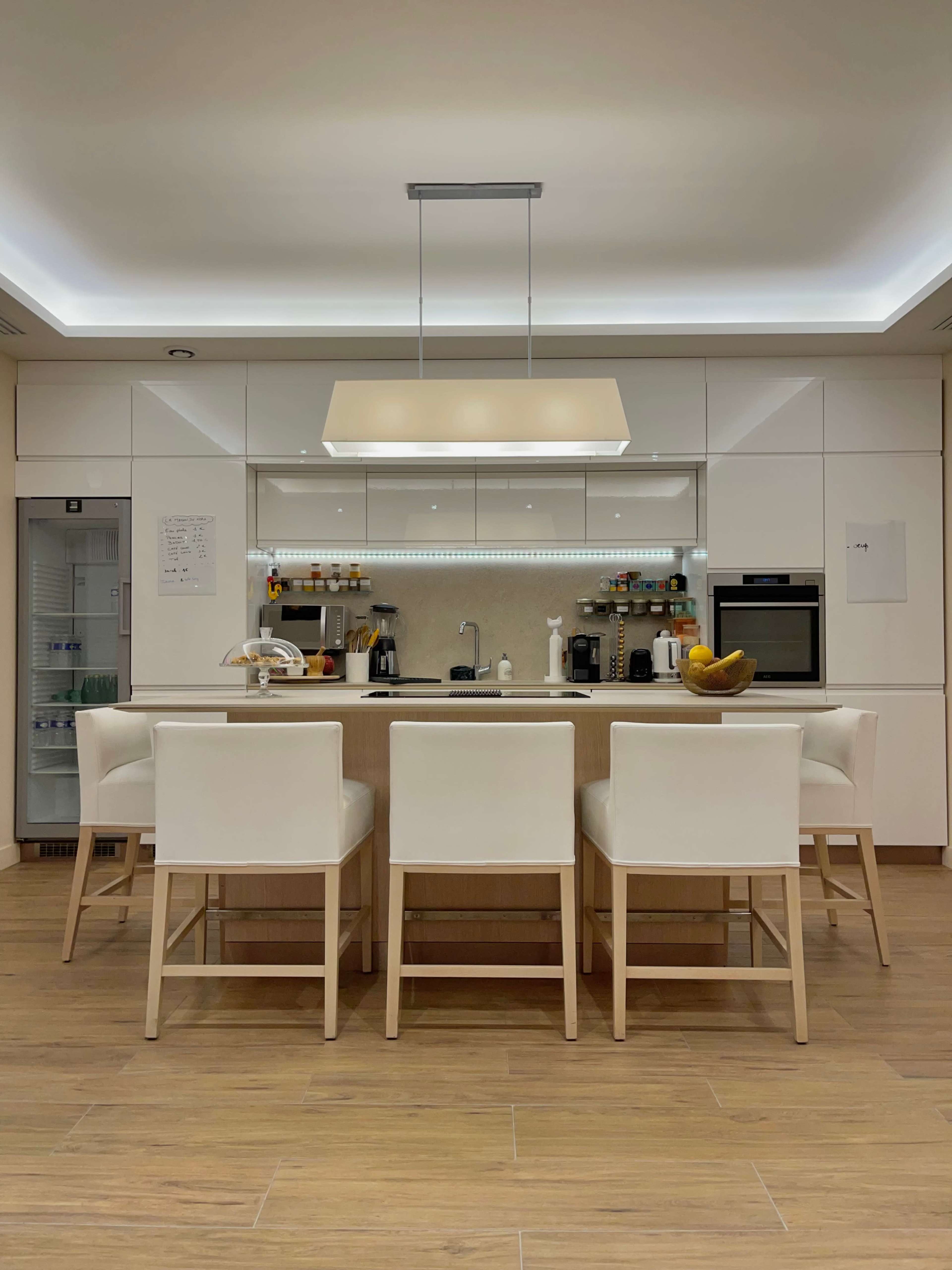 A modern kitchen features a central island with five white bar stools, a light fixture above, and sleek cabinetry lining the walls.
