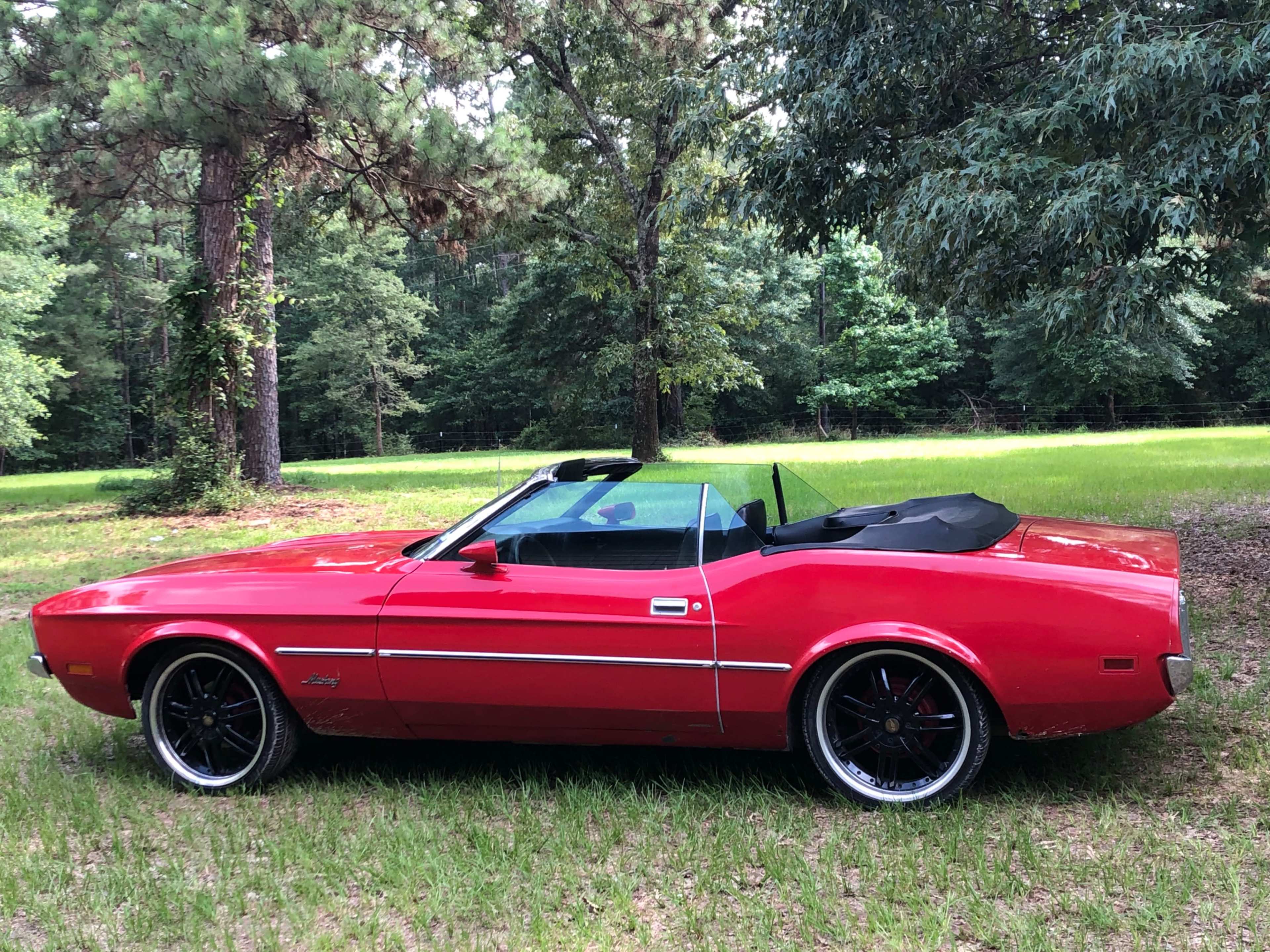 A red convertible car is parked on a grassy area surrounded by trees.
