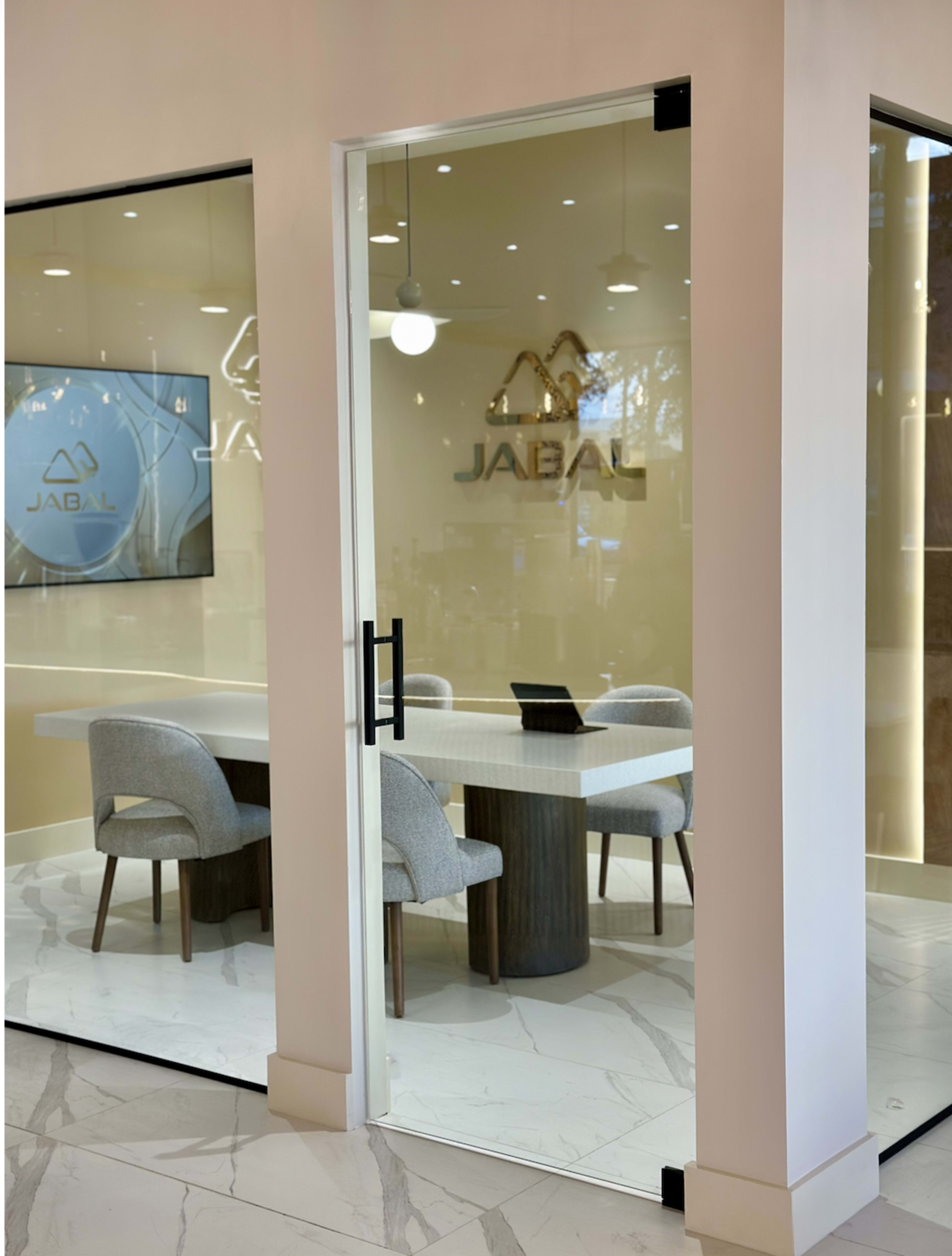 A glass-walled office features a table with four chairs and a large screen displaying the name "JABAL."