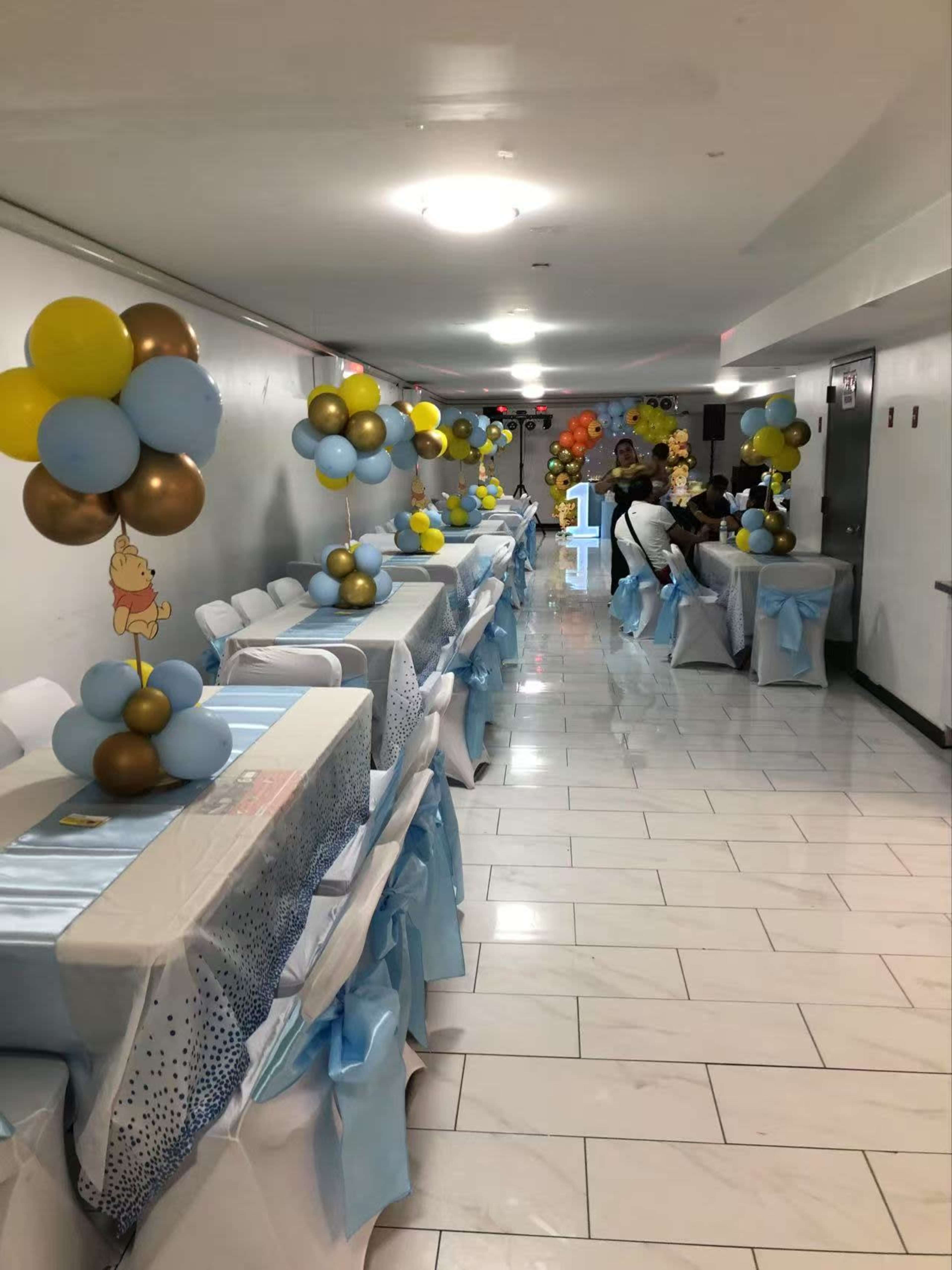 The image shows a decorated hallway with tables covered in light blue and white tablecloths, adorned with colorful balloon centerpieces, and a few attendees seated at the tables.