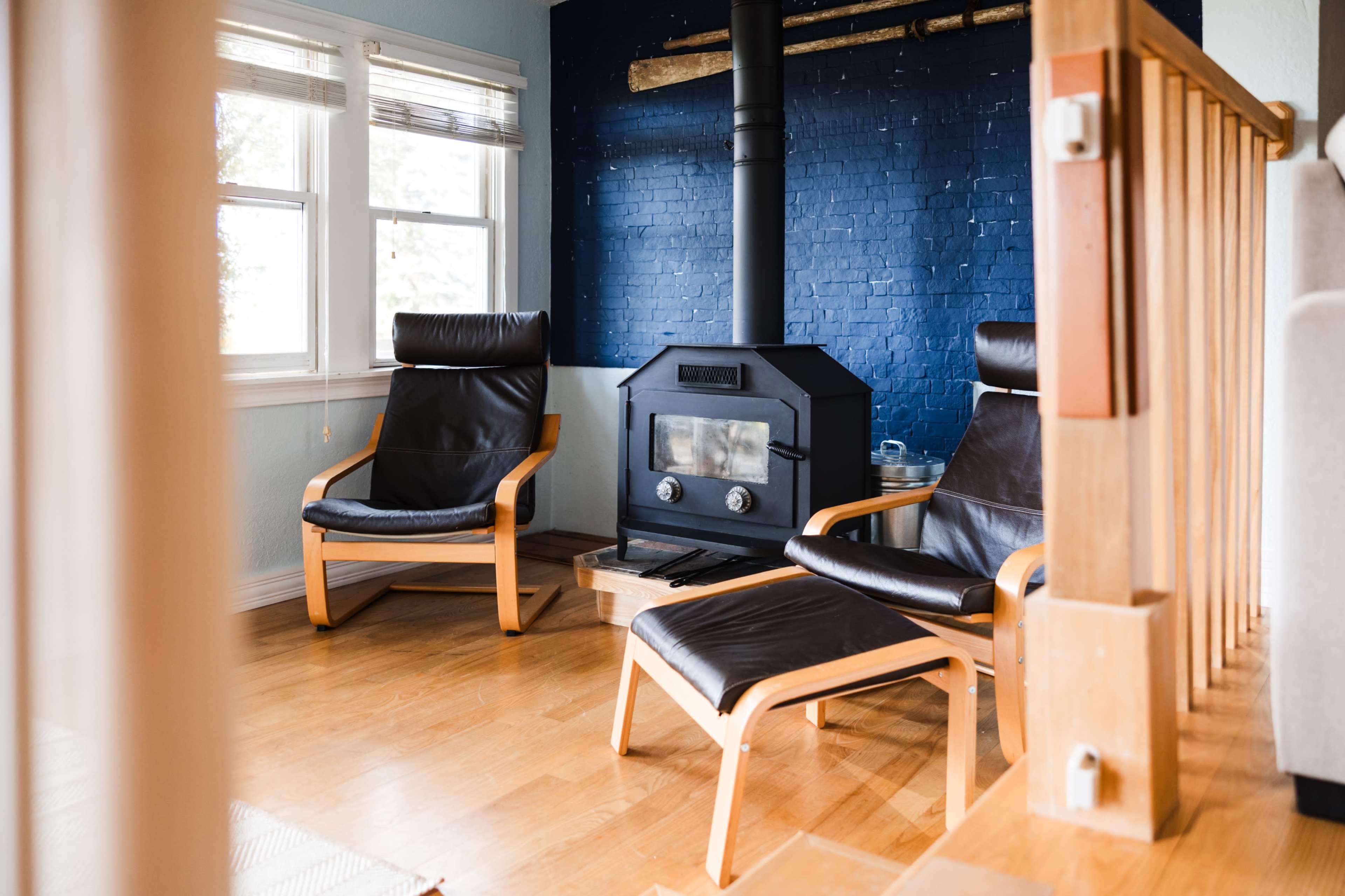 The image shows two black leather chairs positioned near a wood-burning stove in a room with blue brick walls and natural light coming through the windows.