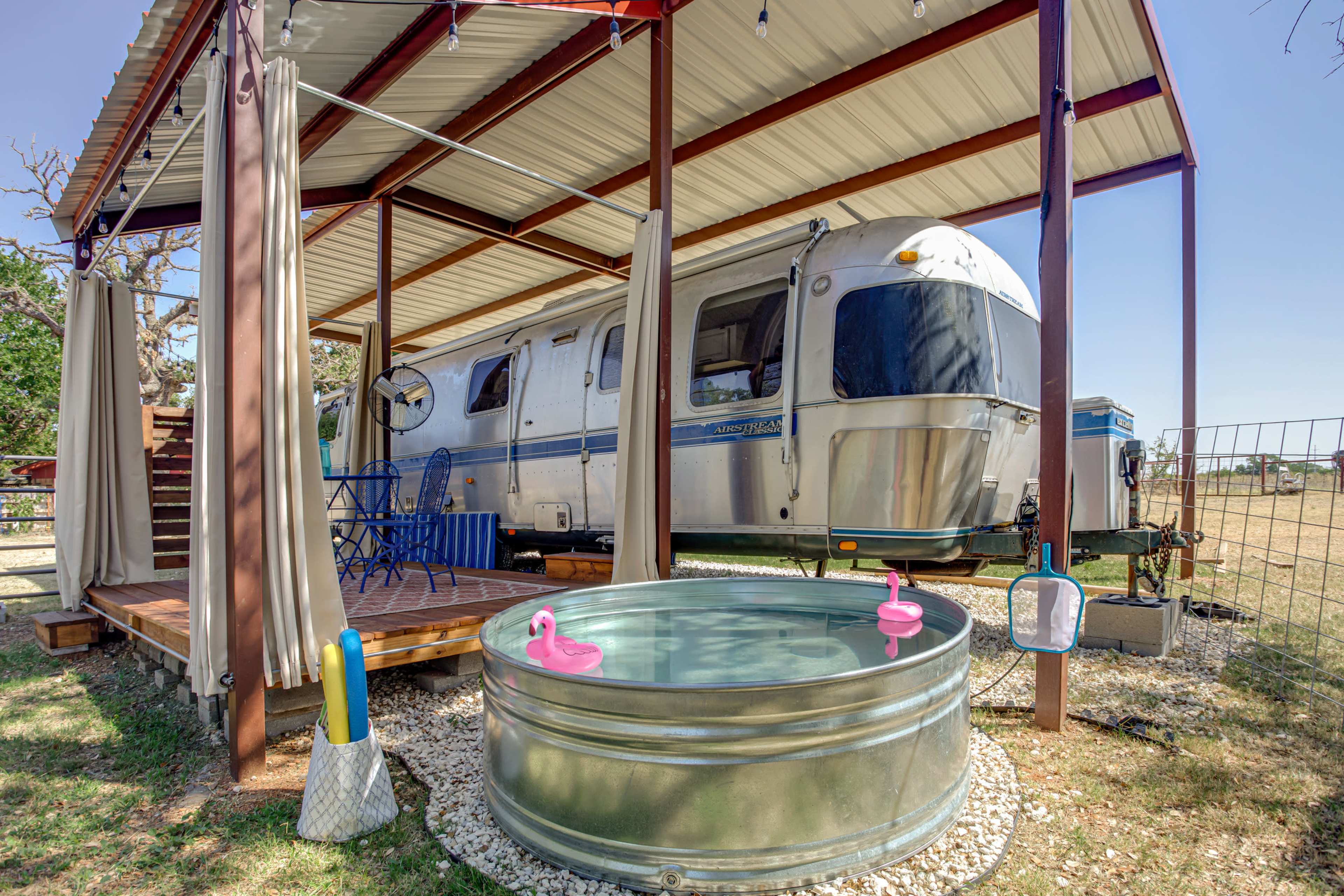 An Airstream trailer is parked under a metal canopy with a wooden deck and a small galvanized tub in front, adorned with pink rubber ducks.